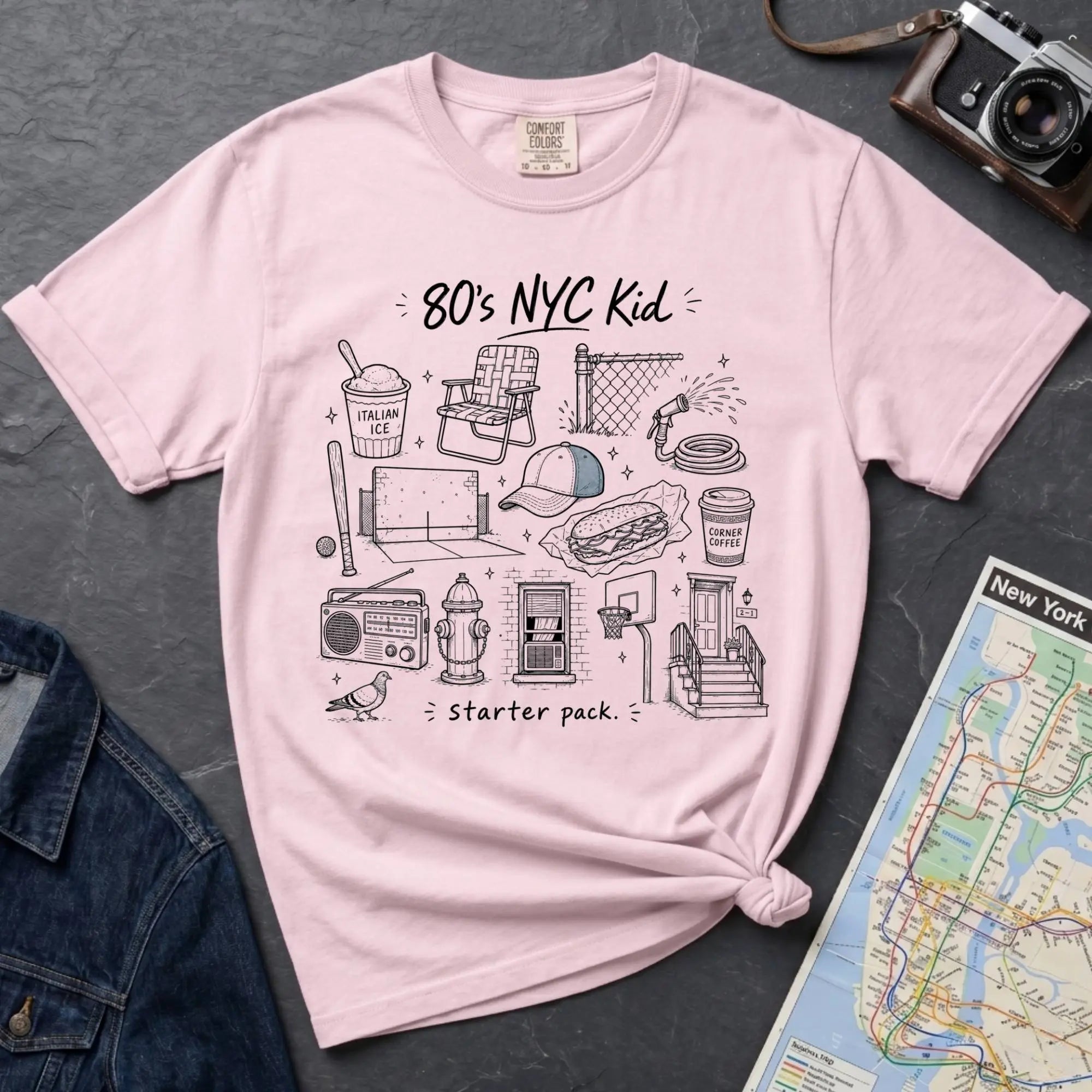 Pink Comfort Colors t-shirt featuring an 80’s NYC Kid Starter Pack graphic with hand-drawn New York City icons including Italian ice, a radio, stoop steps, stickball, a fire hydrant, and a pigeon.