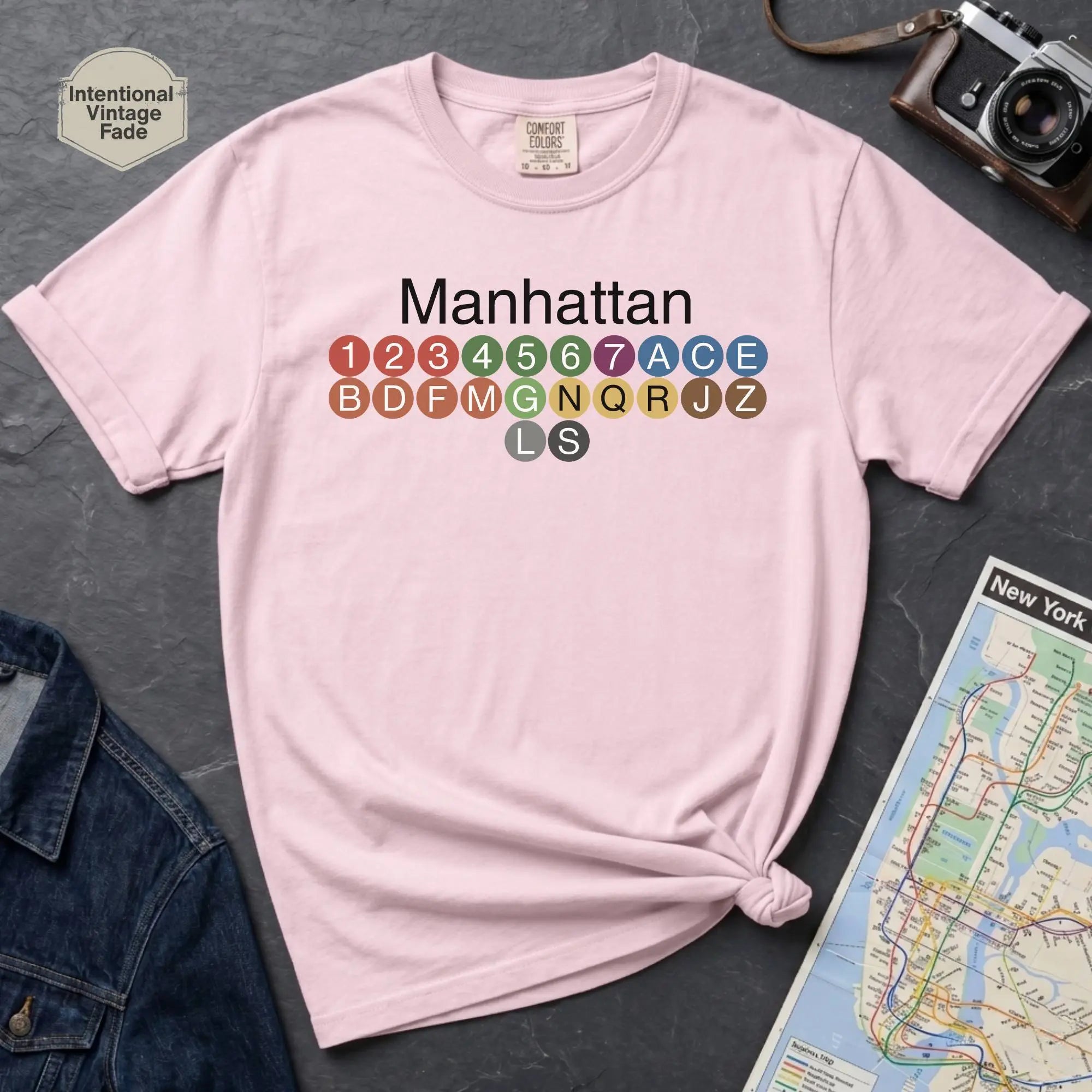 Pink Comfort Colors t-shirt featuring a minimalist Manhattan subway line graphic with colorful NYC train icons.