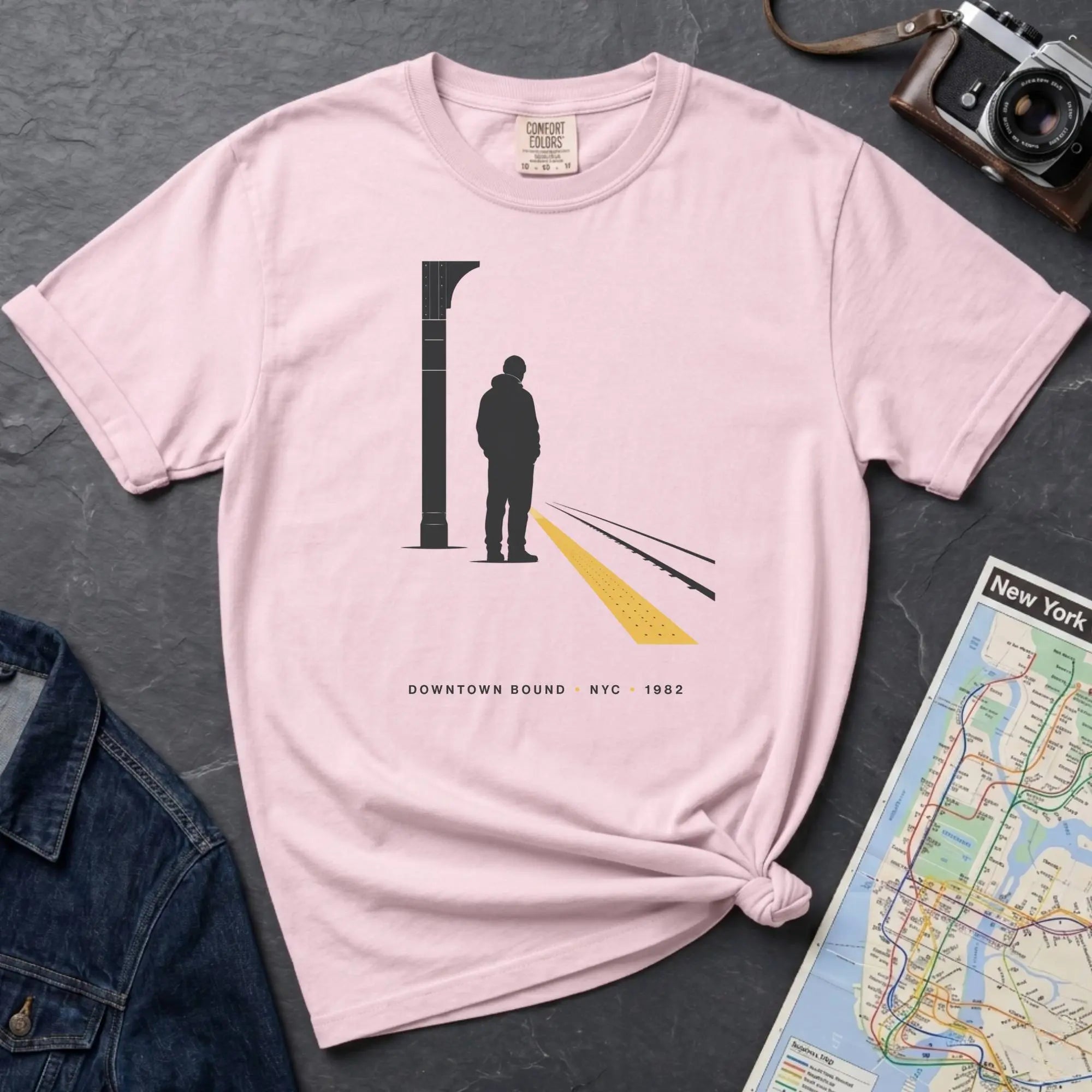 Pink t-shirt with a graphic design of a person on a train platform, placed on a dark surface with a map and denim jacket.