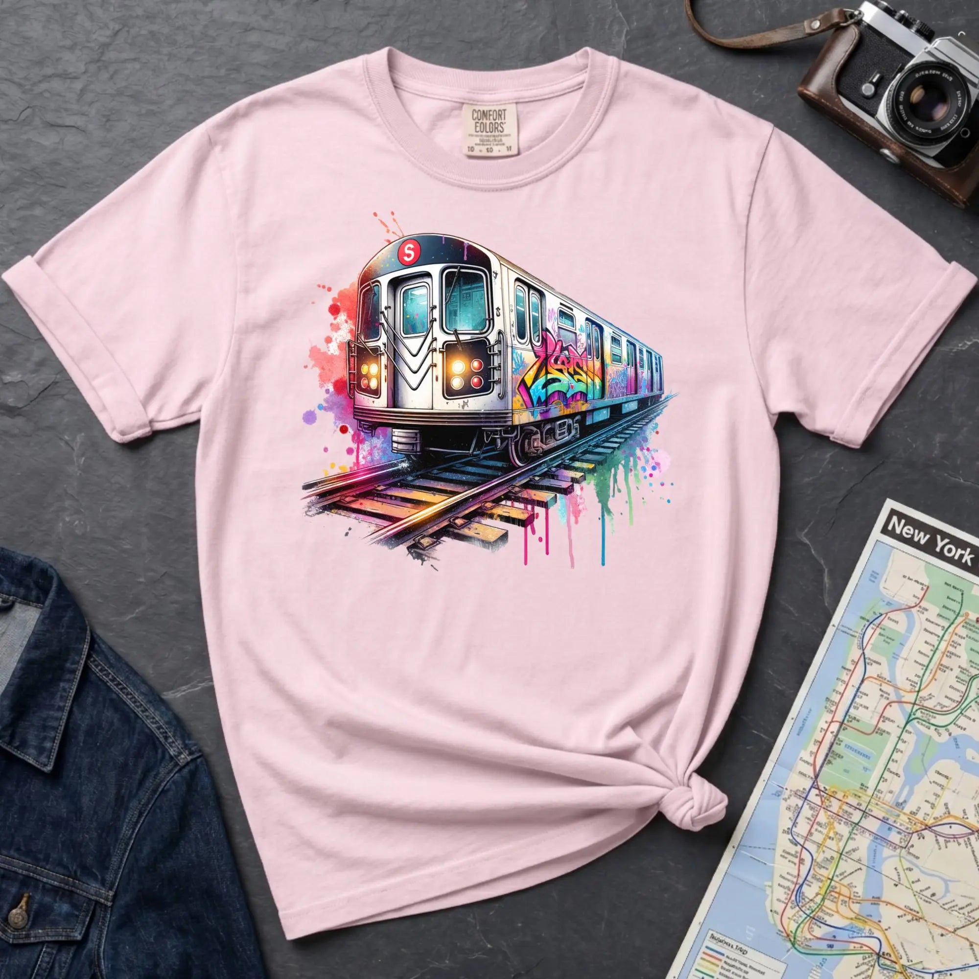 Pink Comfort Colors t-shirt featuring a colorful NYC subway train graphic with graffiti-style paint details and bright street art accents.
