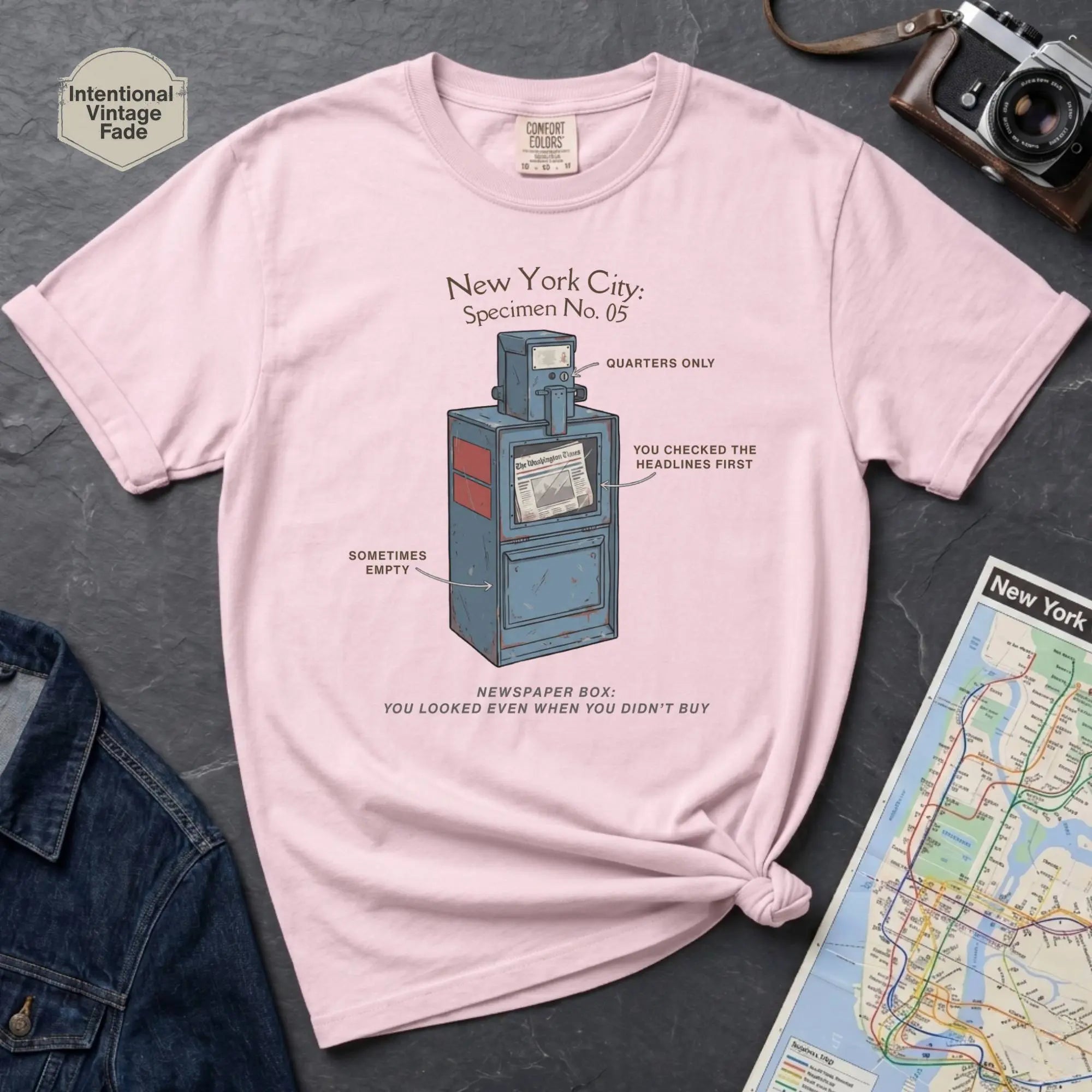 Pink t-shirt with a newspaper box graphic and text on a textured surface with a camera and map.