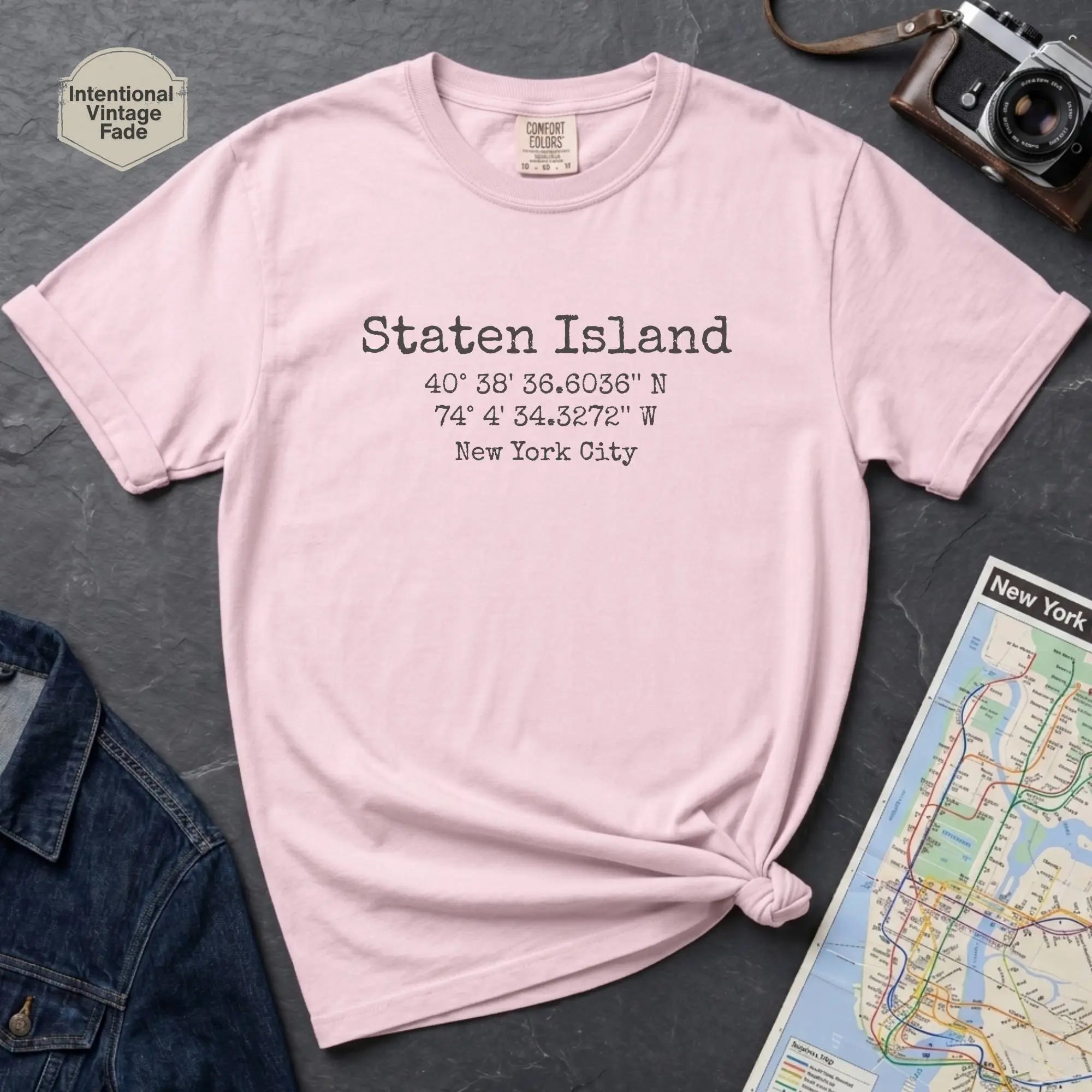 Pink t-shirt with Staten Island text and coordinates on a dark surface with denim jacket and map.