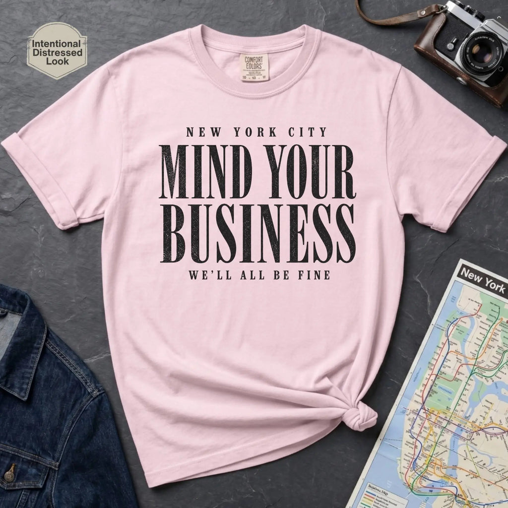 Pink Comfort Colors t-shirt featuring bold New York City “Mind Your Business, We’ll All Be Fine” text in a vintage black typography design.