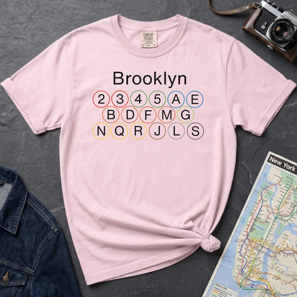 Pink t-shirt with 'Brooklyn' and subway line numbers printed on it, placed on a dark surface with a map and denim jacket.