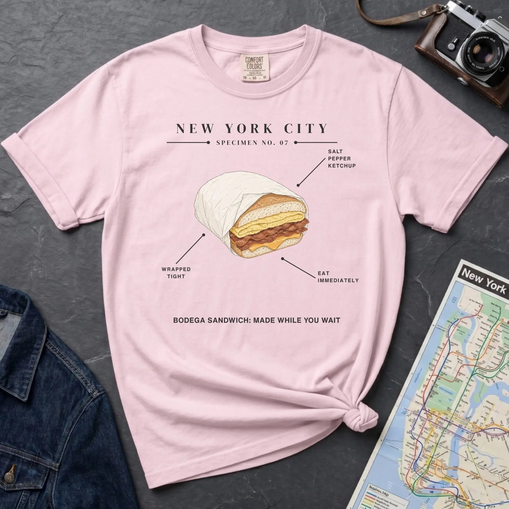 Pink Comfort Colors t-shirt featuring a New York City bodega sandwich graphic with specimen-style callouts and “Bodega Sandwich: Made While You Wait” text.