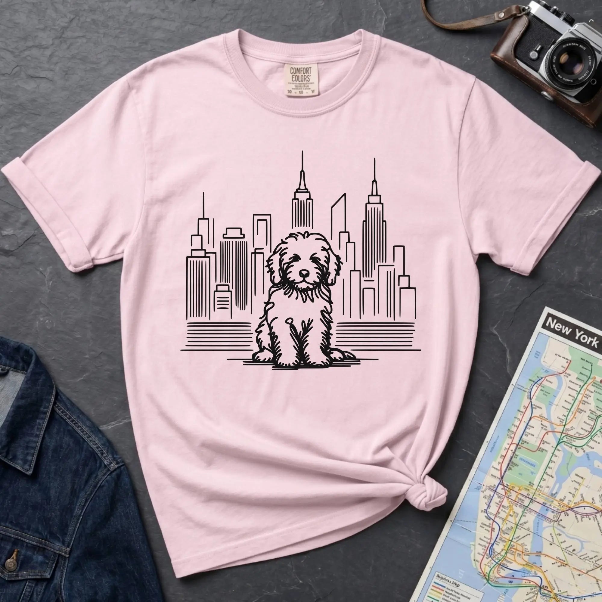 Pink t-shirt with a Golden Doodle dog and cityscape design on a gray surface with a map and camera.