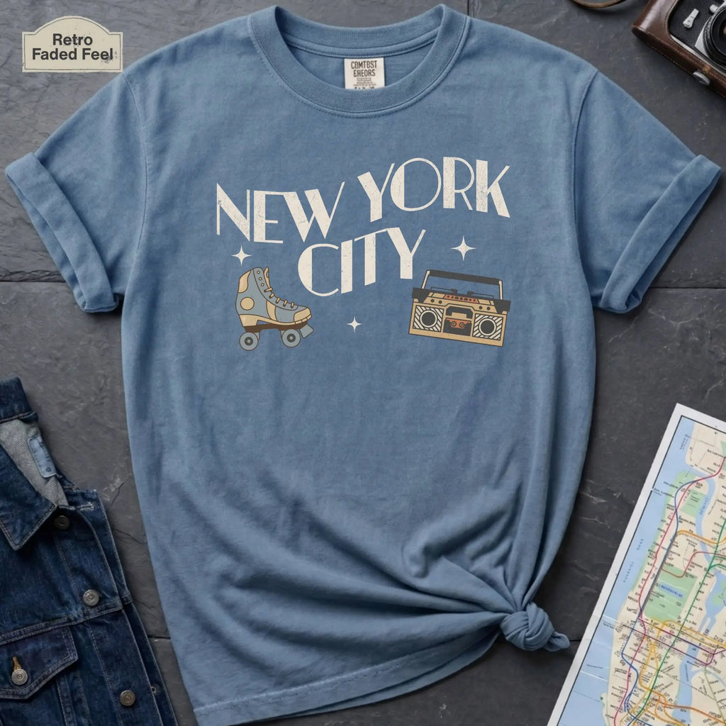 Blue t-shirt with 'New York City' text and a roller skate and boom box on a dark surface with a map and camera.