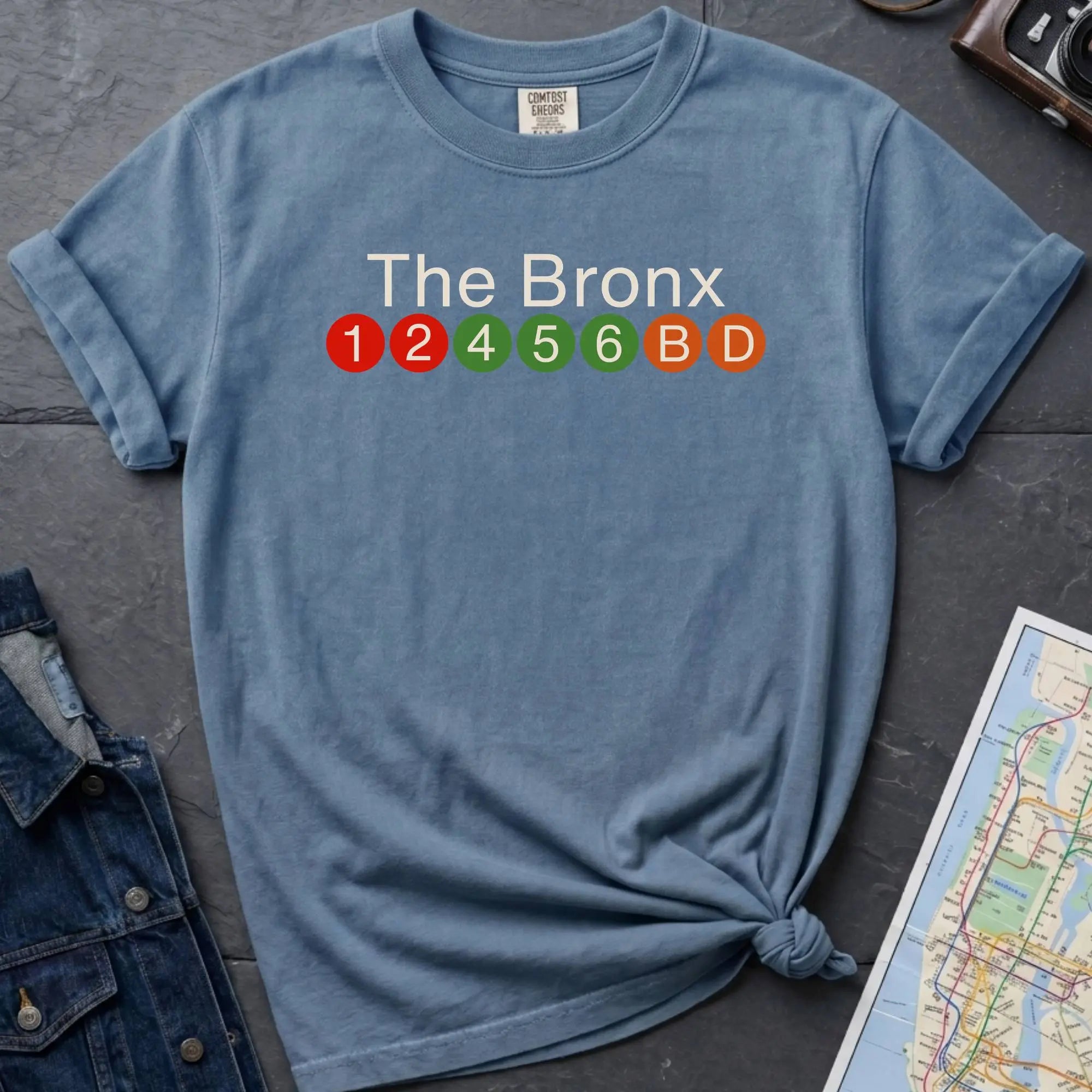 Blue Comfort Colors t-shirt featuring a minimalist Bronx subway line graphic with The Bronx text and colorful NYC train icons.
