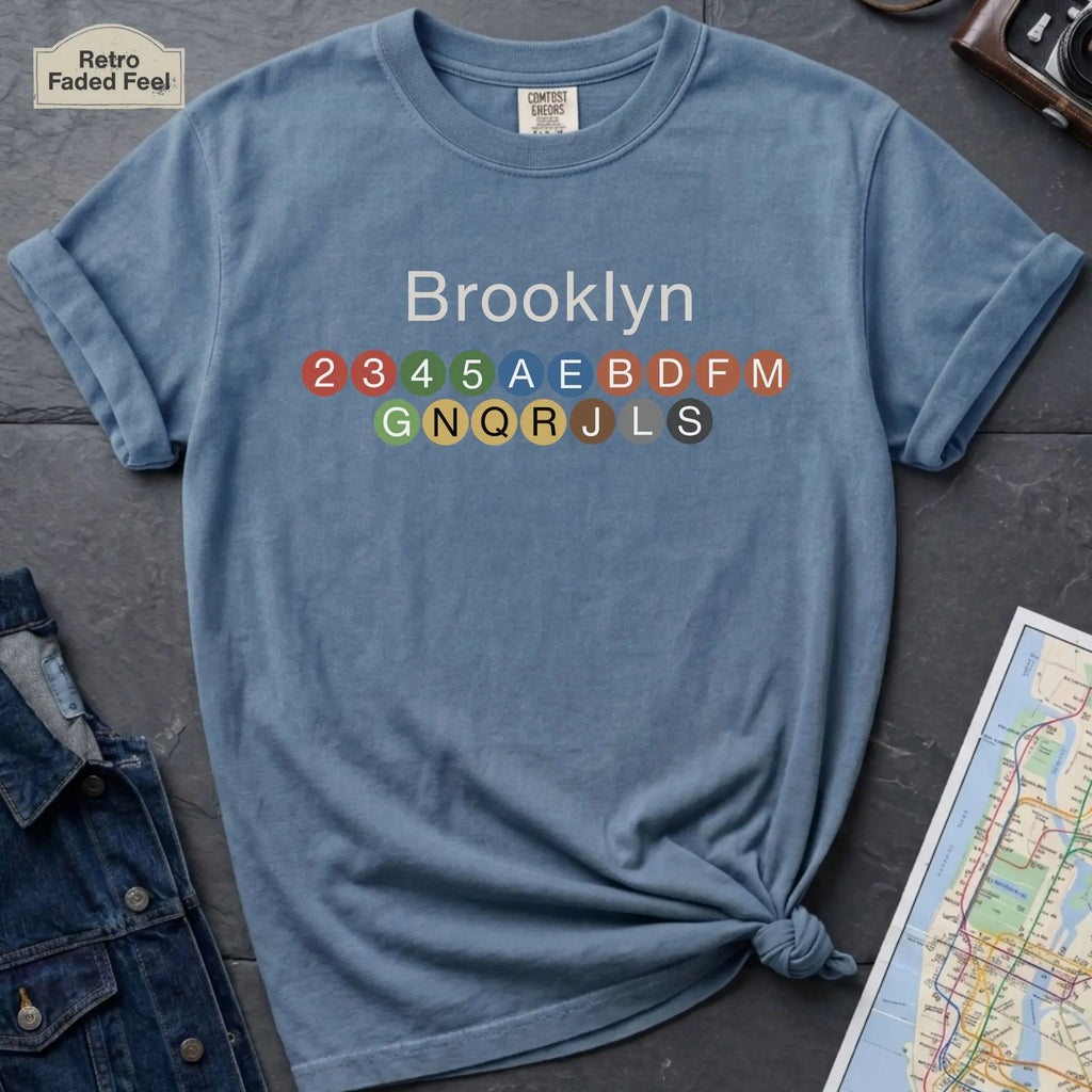 Blue t-shirt with 'Brooklyn' and subway line numbers on a dark surface with a map and camera.