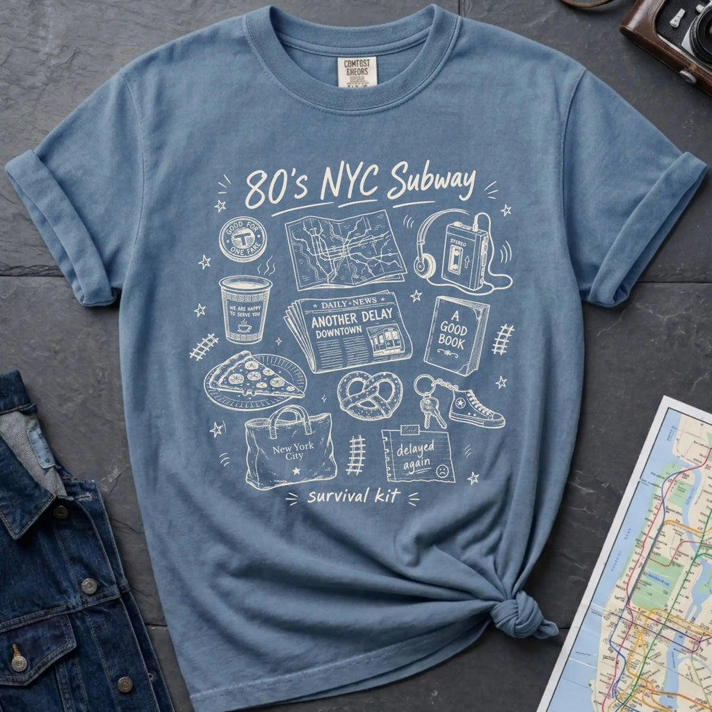 Blue t-shirt with '80s NYC Subway Survival Kit' graphic design on a dark surface with a map and denim jacket.