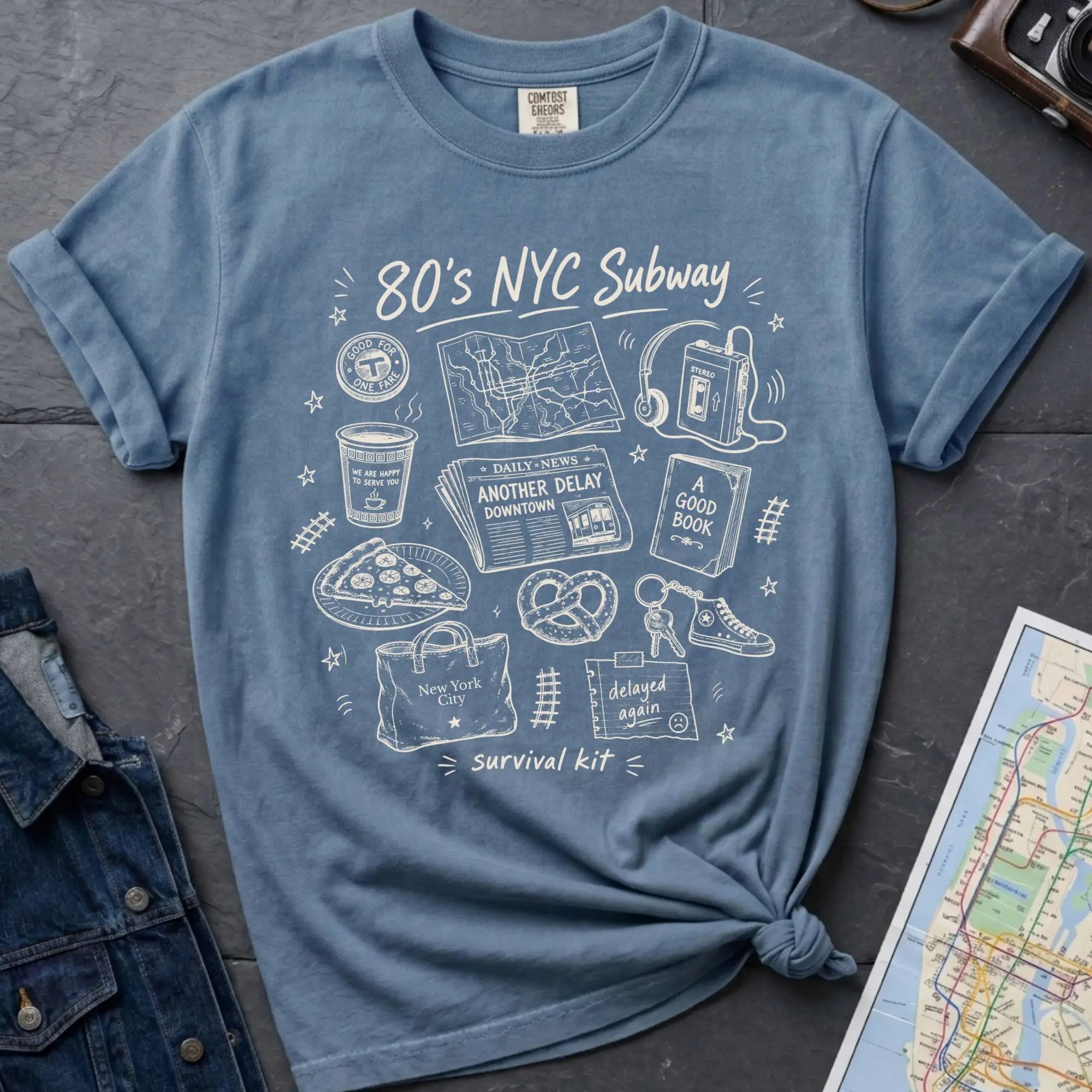 Blue t-shirt with '80s NYC Subway Survival Kit' graphic design on a dark surface with a map and denim jacket.