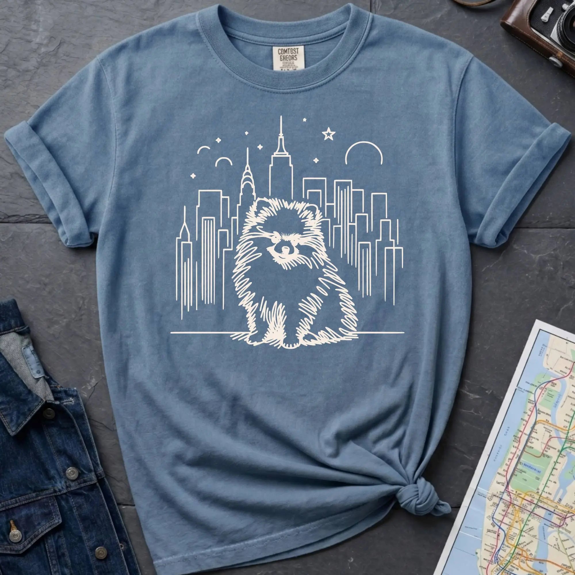 Blue Comfort Colors t-shirt featuring a fluffy small dog illustration in front of the New York City skyline.