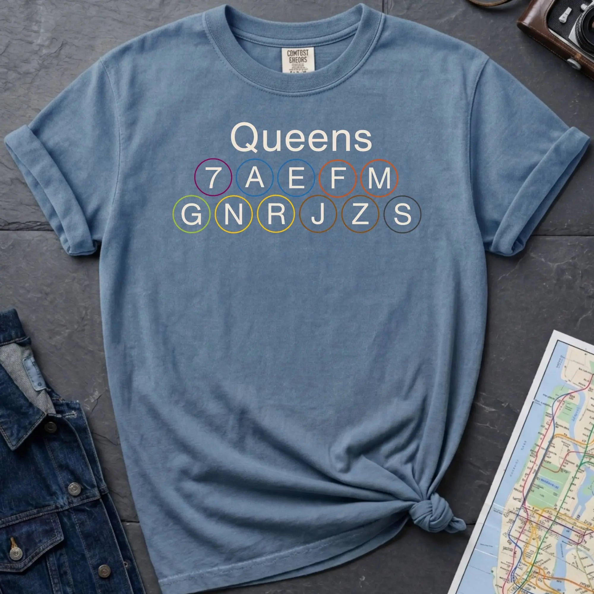 Blue Comfort Colors t-shirt featuring a minimalist Queens subway line graphic with Queens text and classic NYC train icons.