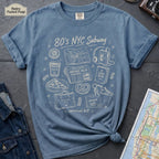 Blue t-shirt with '80s NYC Subway Survival Kit' graphic on a dark surface with a map and jeans.