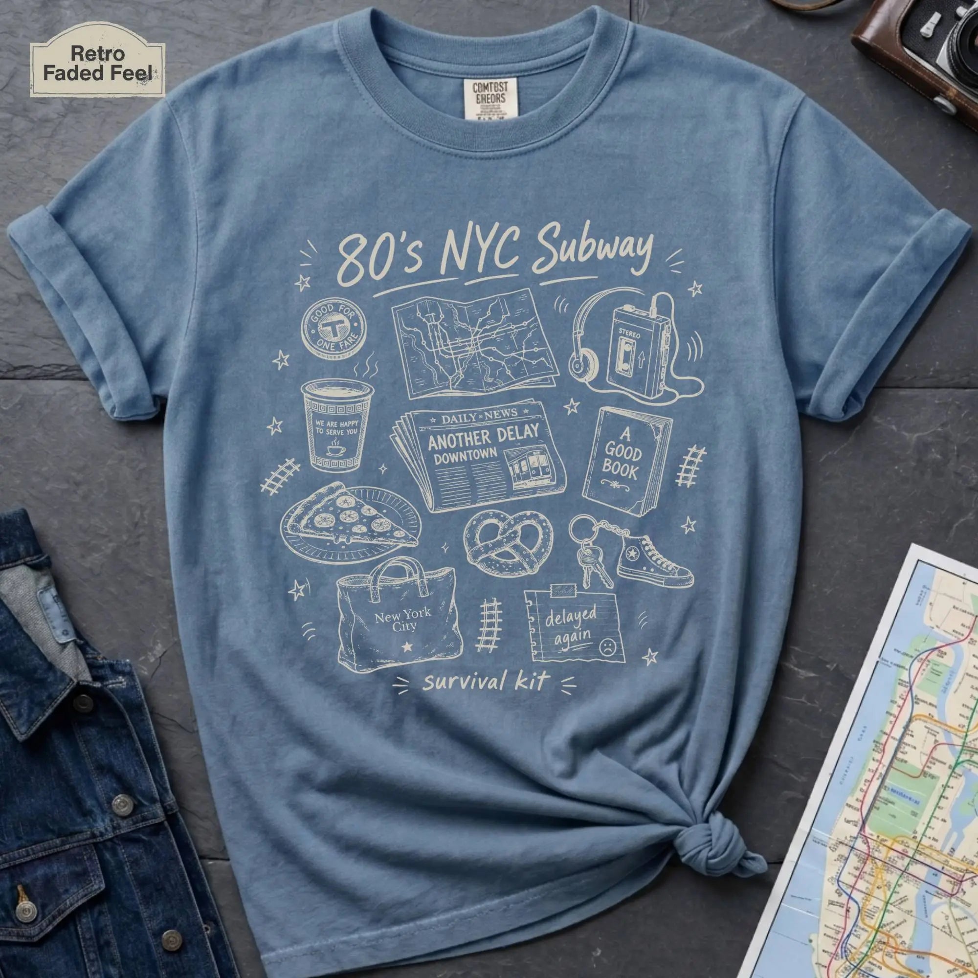 Blue t-shirt with '80s NYC Subway Survival Kit' graphic on a dark surface with a map and jeans.