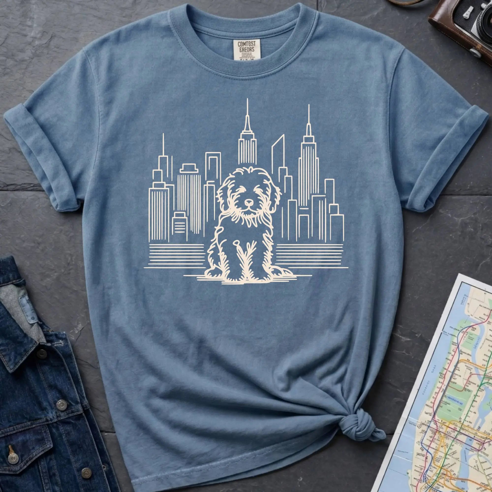 Blue t-shirt with a Golden Doodle and cityscape design on a dark surface with a map and camera.