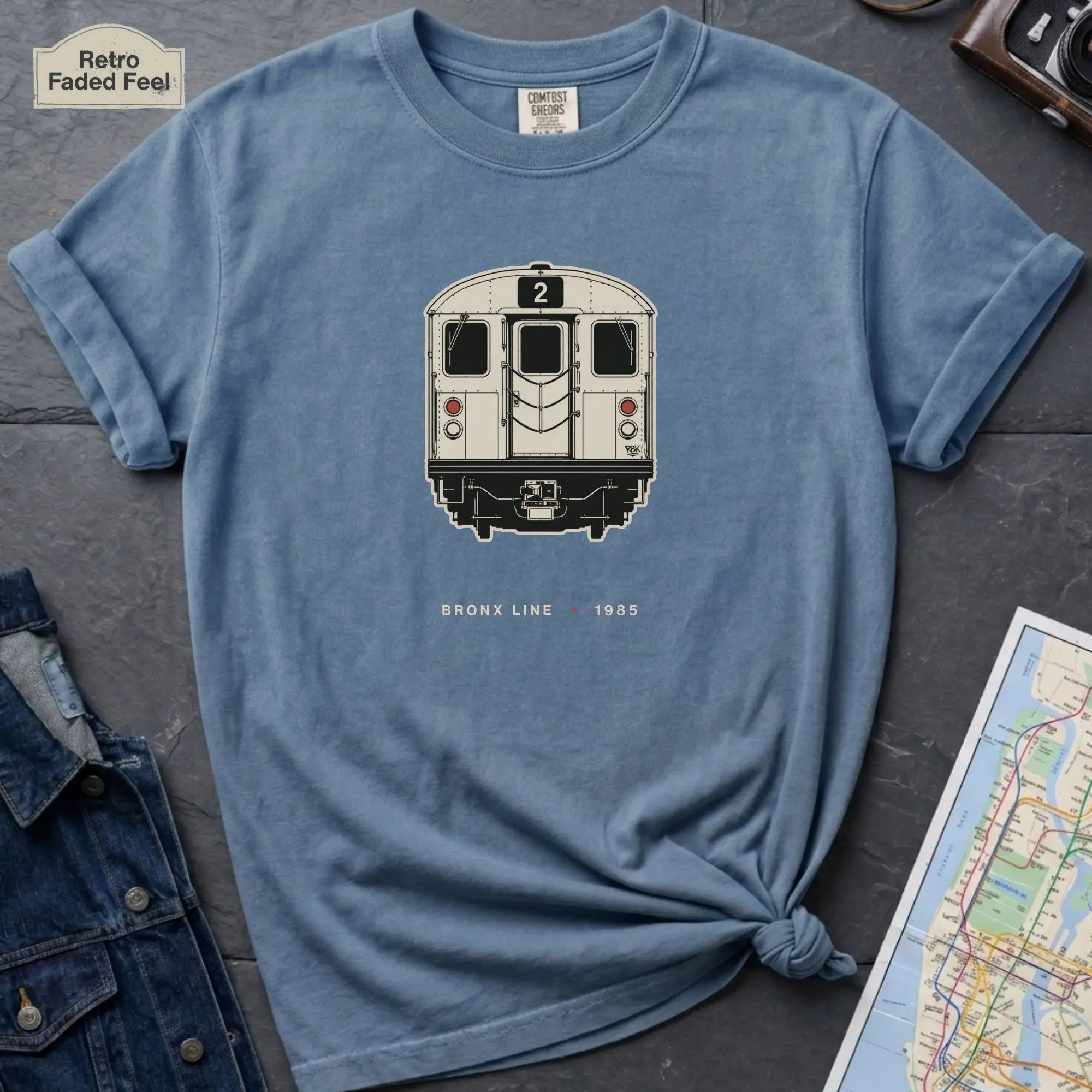 Blue t-shirt with a NYC subway train graphic on a dark surface with a map and a denim jacket.