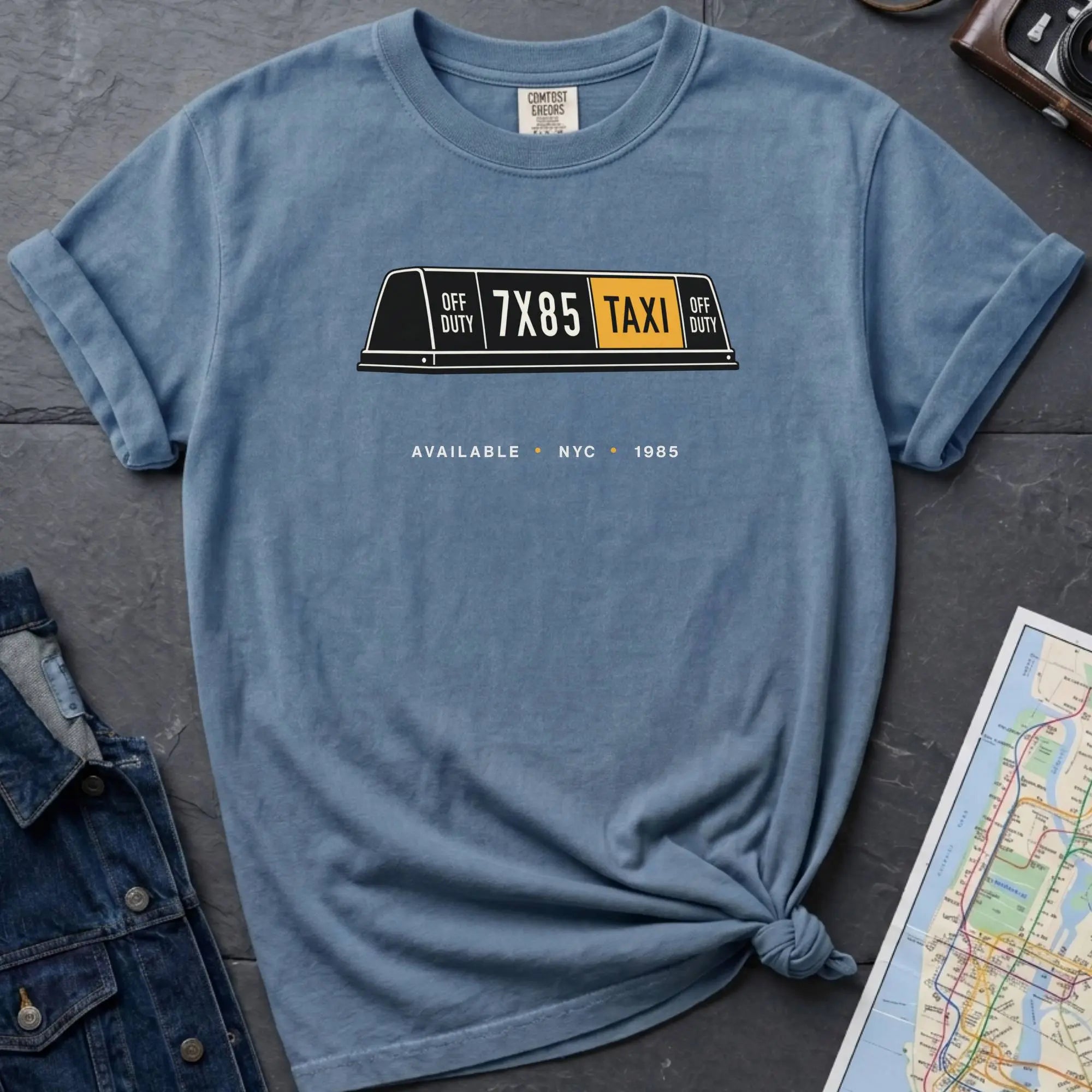 Blue Comfort Colors t-shirt featuring a vintage New York City taxi roof sign graphic with “Available • NYC • 1985” text.