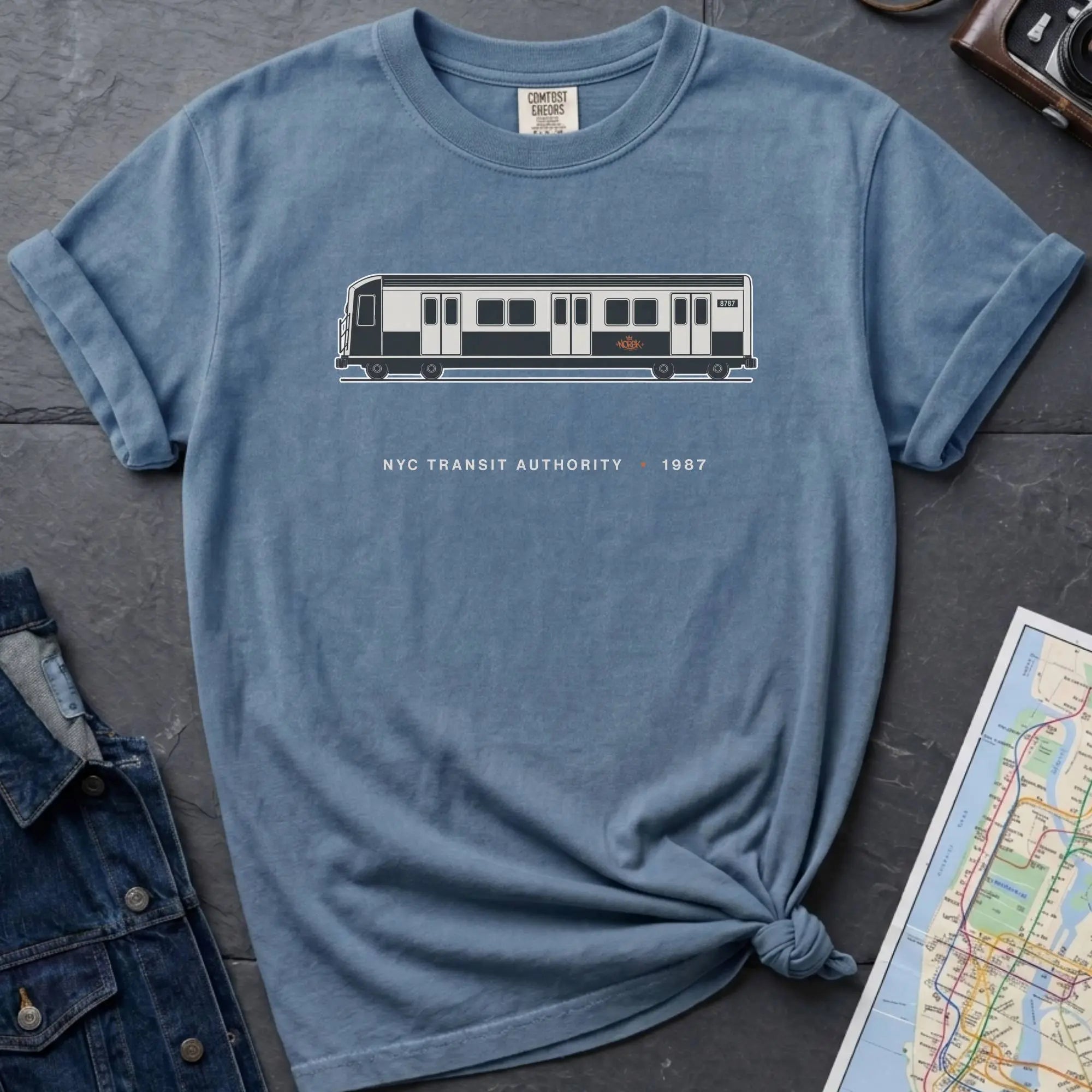 Blue Comfort Colors t-shirt featuring a minimalist New York City subway car graphic with “NYC Transit Authority • 1987” text.