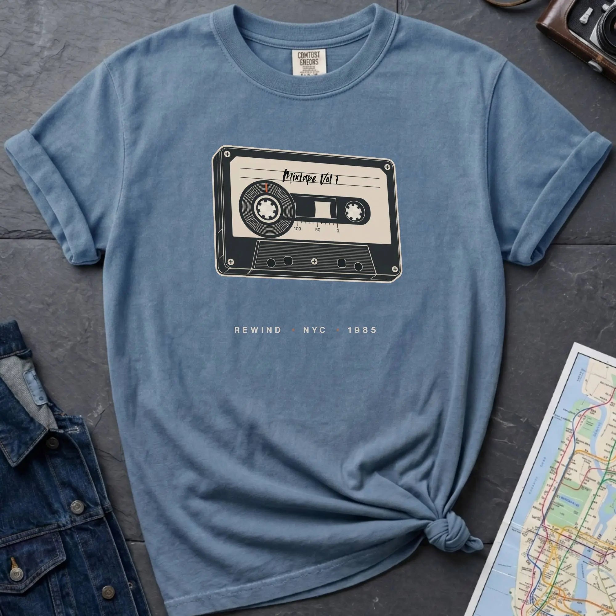 Blue t-shirt with cassette tape graphic and text on a gray surface with denim jacket and map.