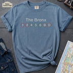 Blue t-shirt with 'The Bronx' and subway line numbers on a dark surface with a map and camera.