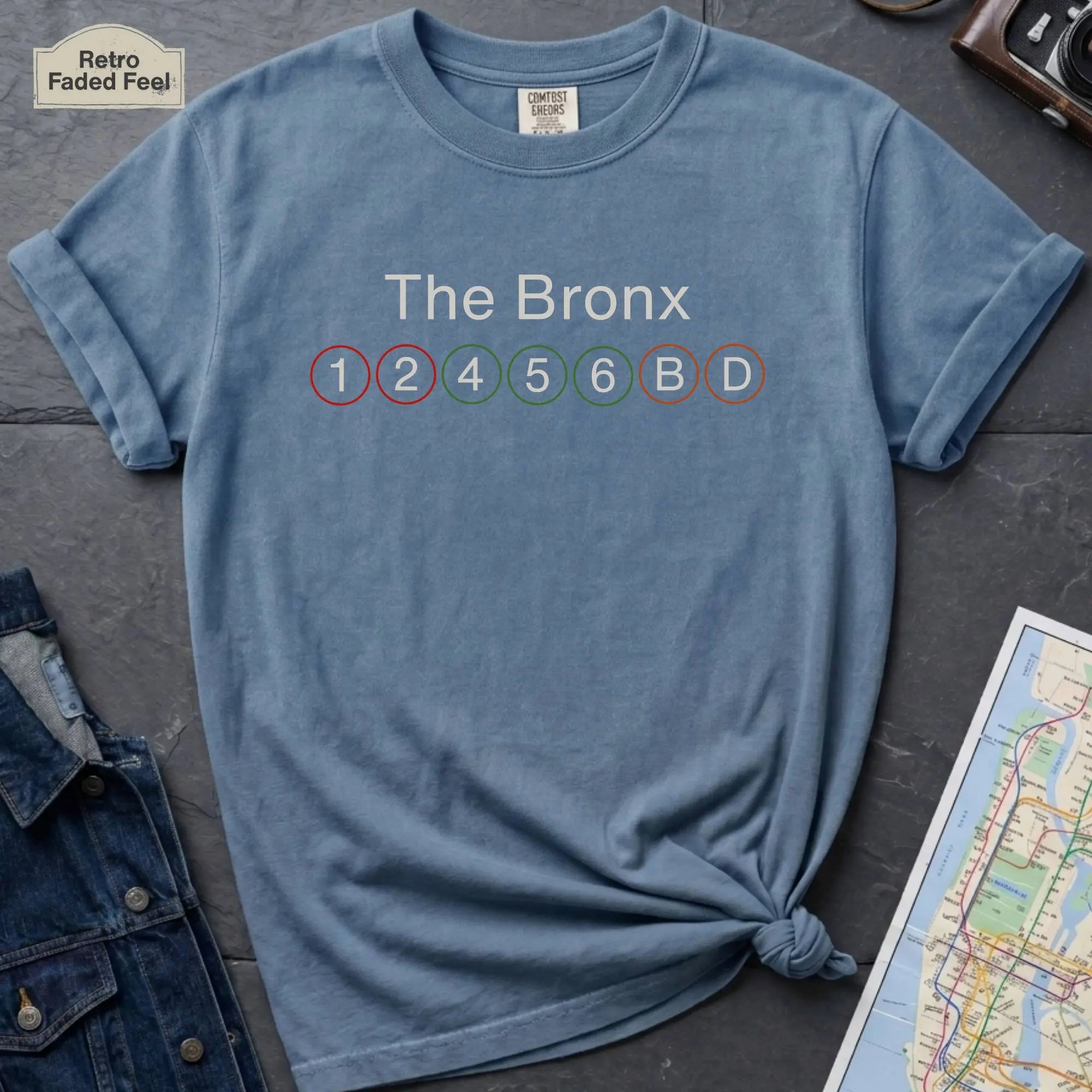 Blue t-shirt with 'The Bronx' and subway line numbers on a dark surface with a map and camera.