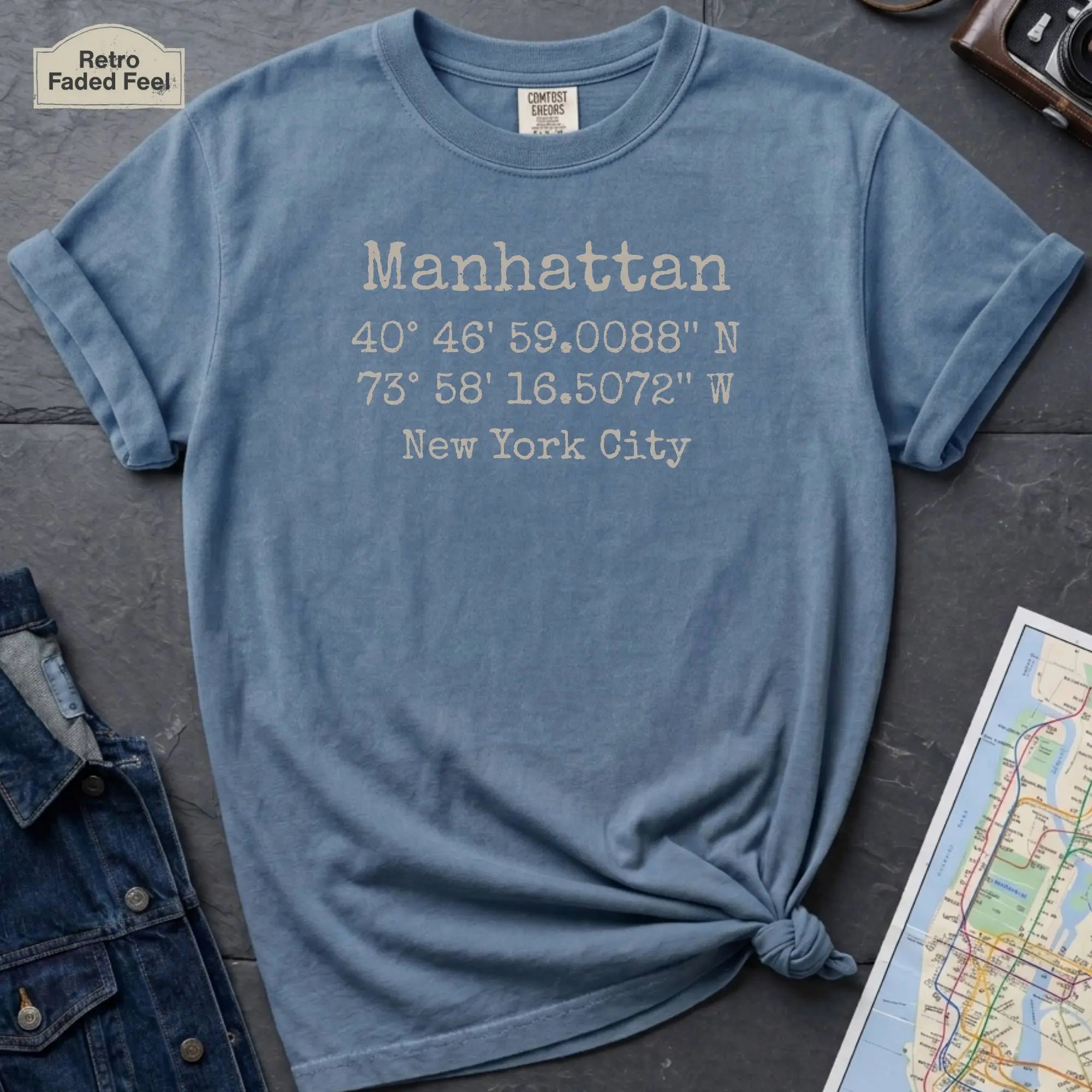 Blue Comfort Colors t-shirt featuring Manhattan, NY coordinates and New York City text in a minimalist typewriter-style design.