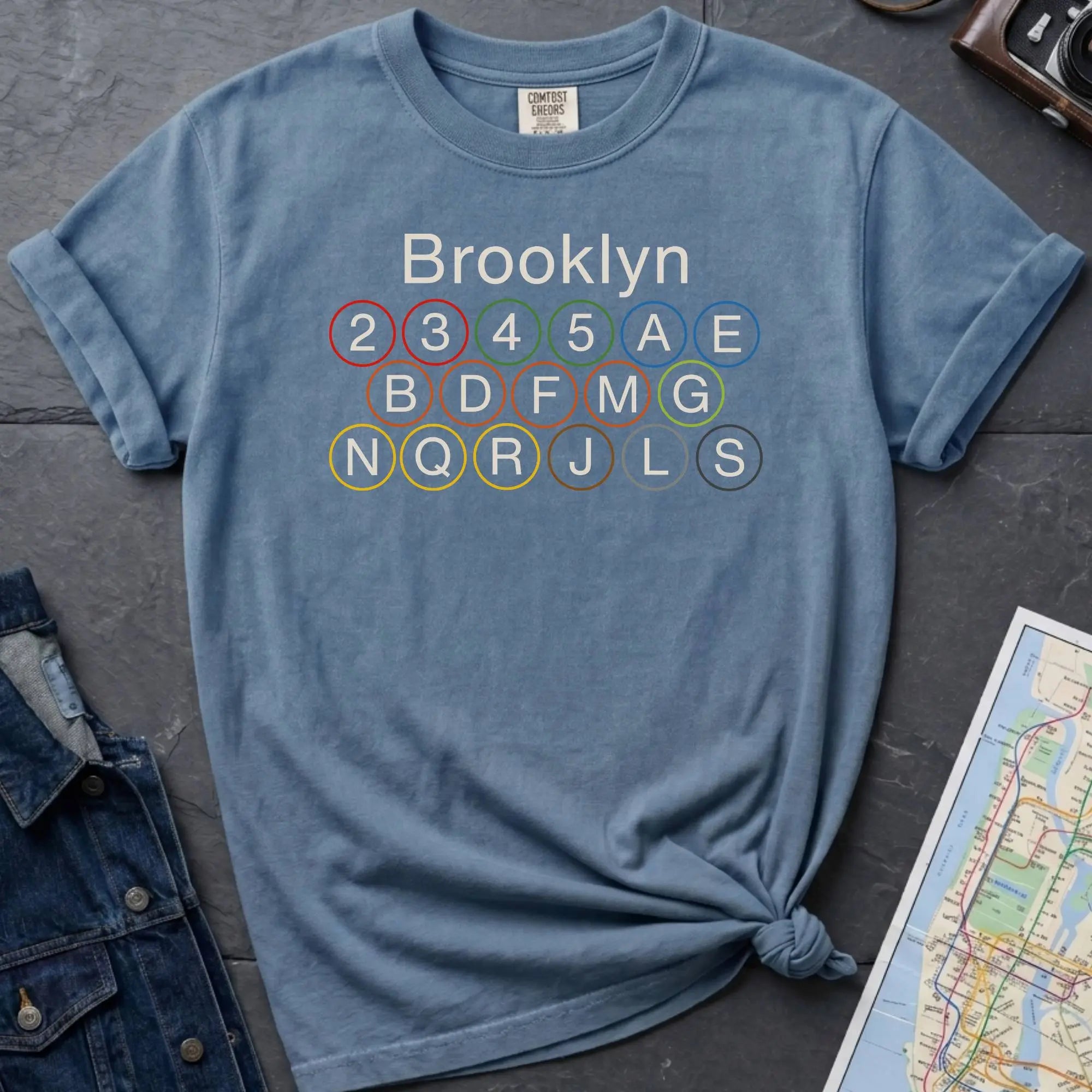 Blue Comfort Colors t-shirt featuring the word “Brooklyn” above colorful NYC subway line circles representing the subway routes that run through Brooklyn.