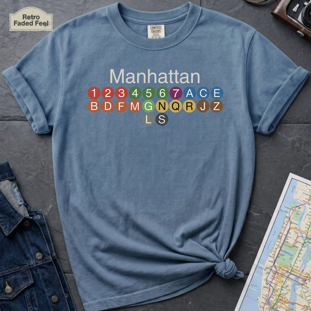 Blue Comfort Colors t-shirt featuring a minimalist Manhattan subway line graphic with colorful NYC train icons.