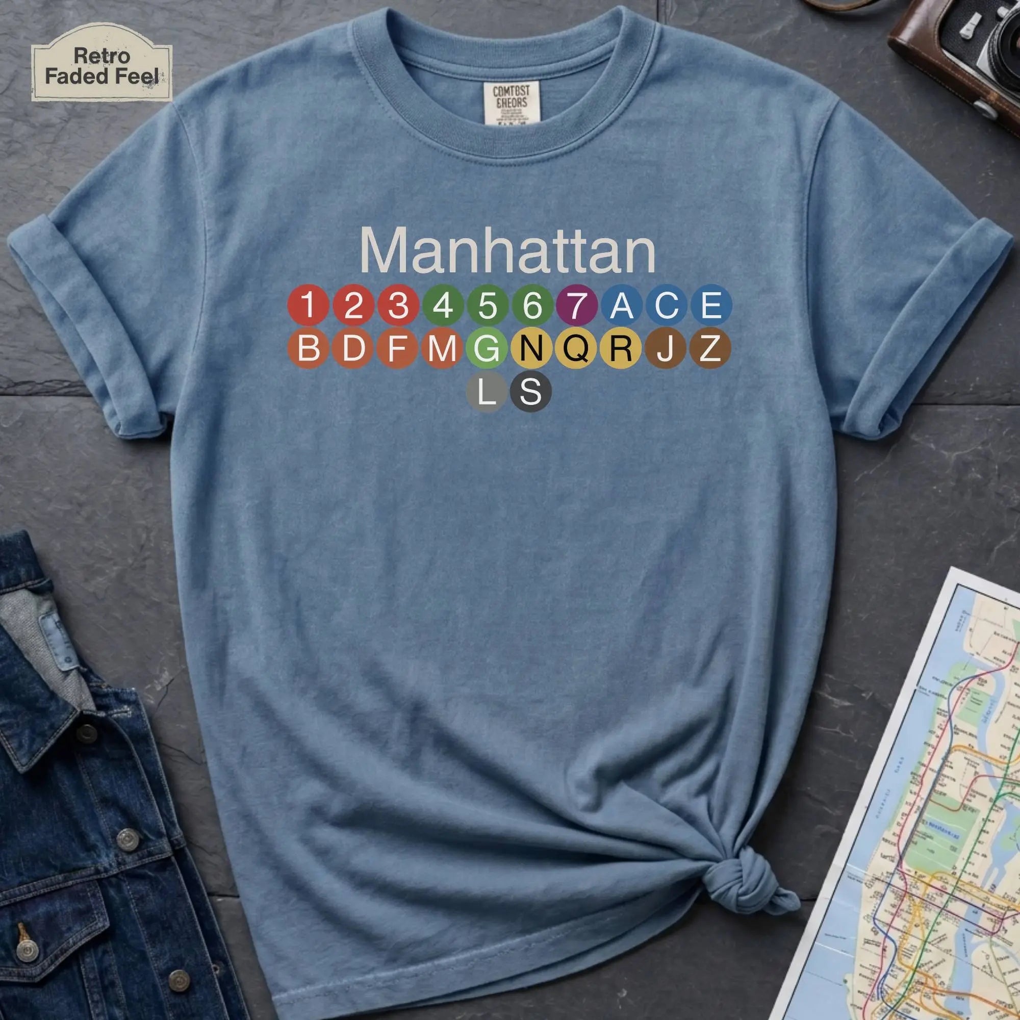 Blue Comfort Colors t-shirt featuring a minimalist Manhattan subway line graphic with colorful NYC train icons.