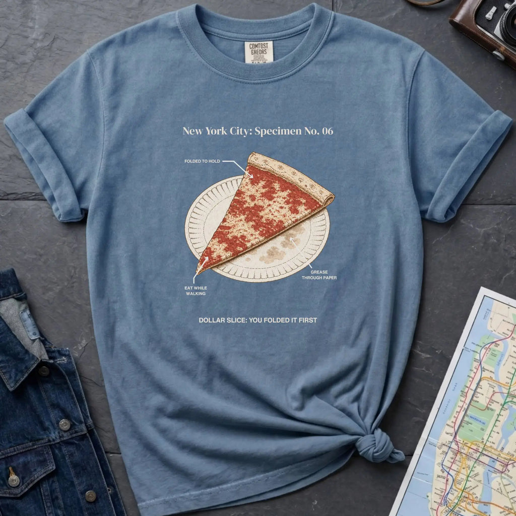 Blue Comfort Colors t-shirt featuring a vintage New York City dollar slice pizza graphic with specimen-style callouts and a folded slice on a paper plate.