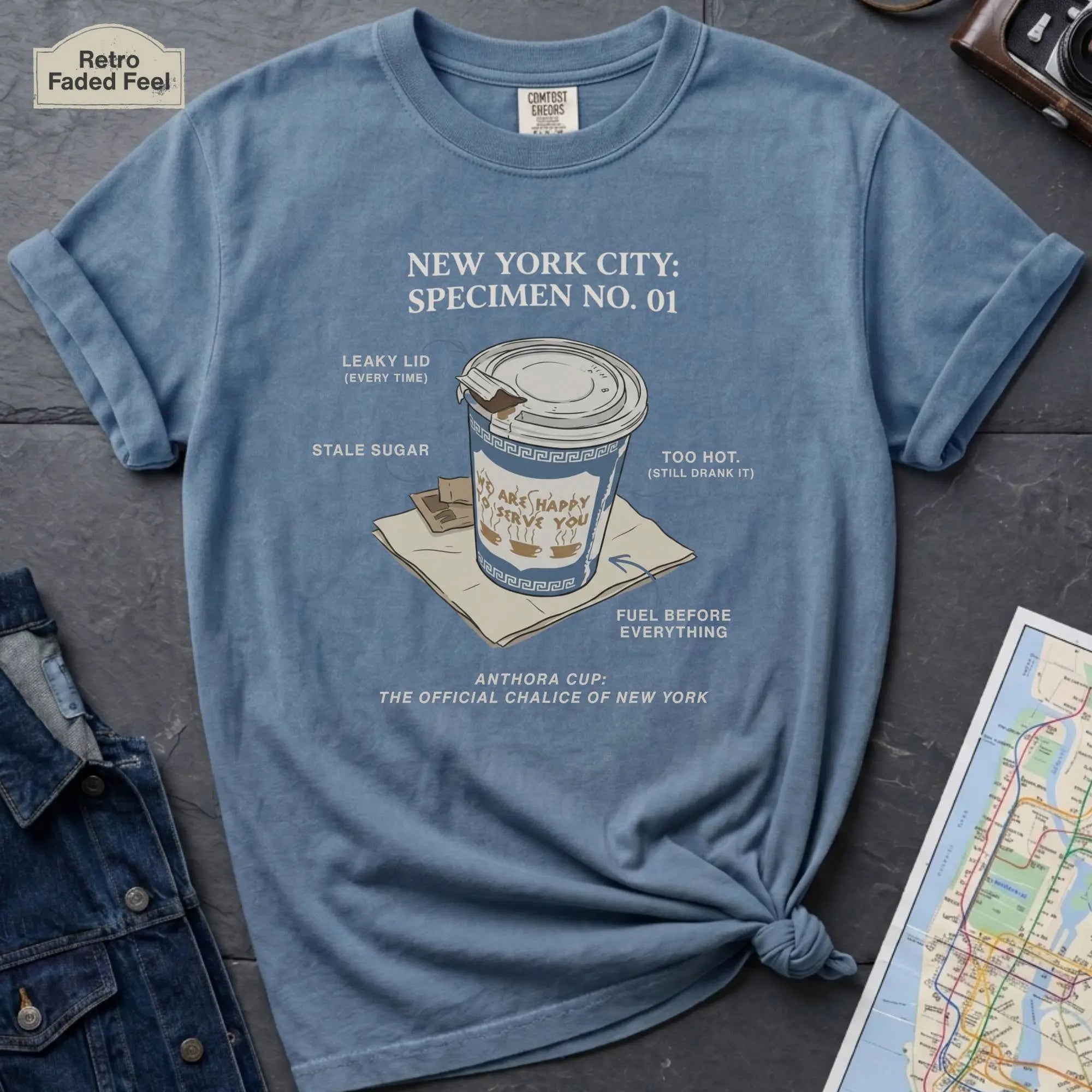 Blue t-shirt with New York City coffee cup graphic on a textured surface with map and camera.