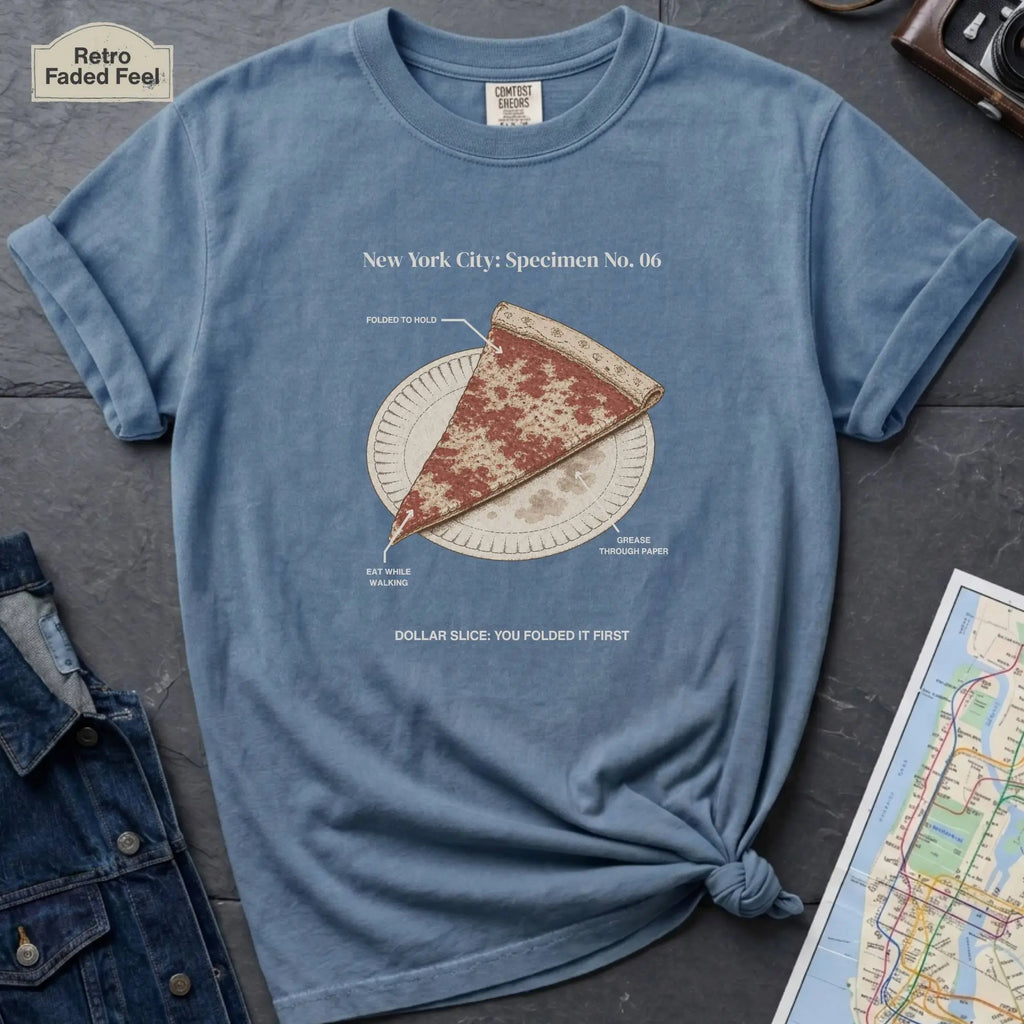 Blue Comfort Colors t-shirt featuring a vintage New York City dollar slice pizza graphic with specimen-style callouts and a folded slice on a paper plate.