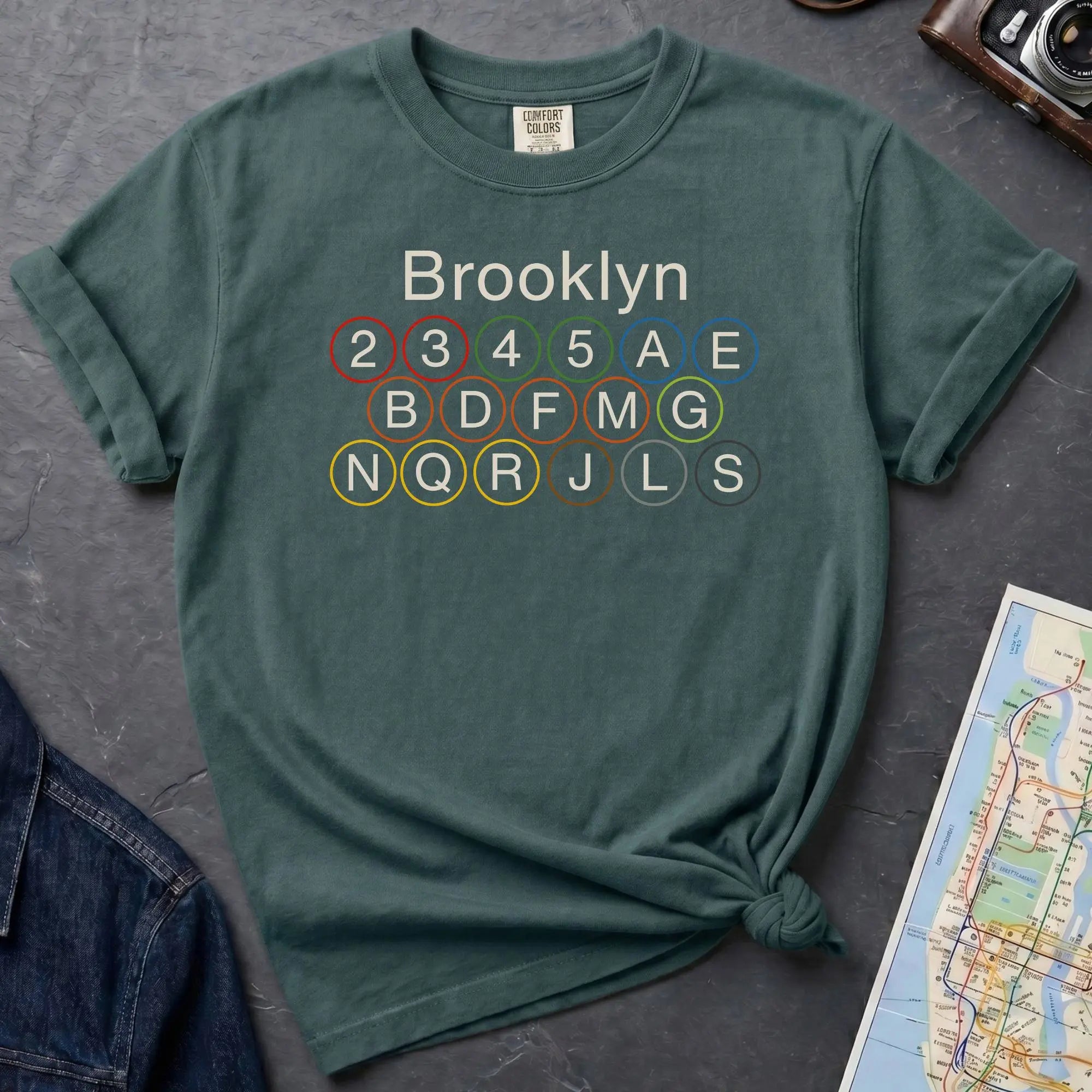 Blue Spruce Comfort Colors t-shirt featuring the word “Brooklyn” above colorful NYC subway line circles representing the subway routes that run through Brooklyn.