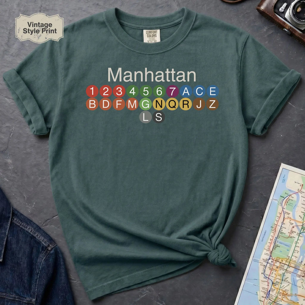 Blue Spruce Comfort Colors t-shirt featuring a minimalist Manhattan subway line graphic with colorful NYC train icons.