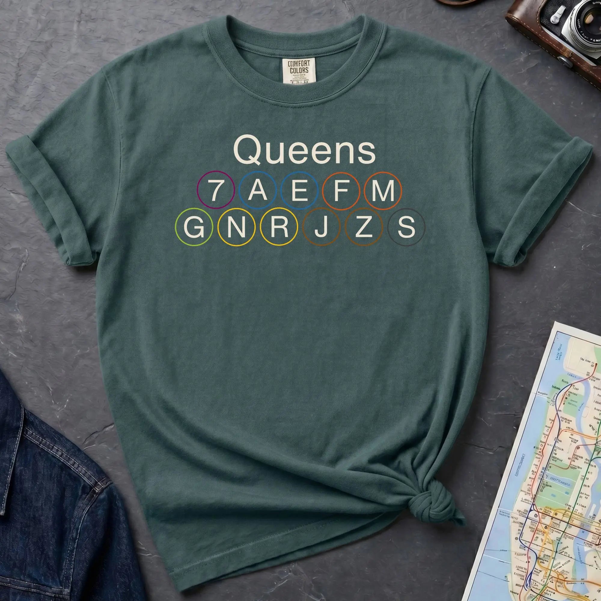 Blue Spruce Comfort Colors t-shirt featuring a minimalist Queens subway line graphic with Queens text and classic NYC train icons.