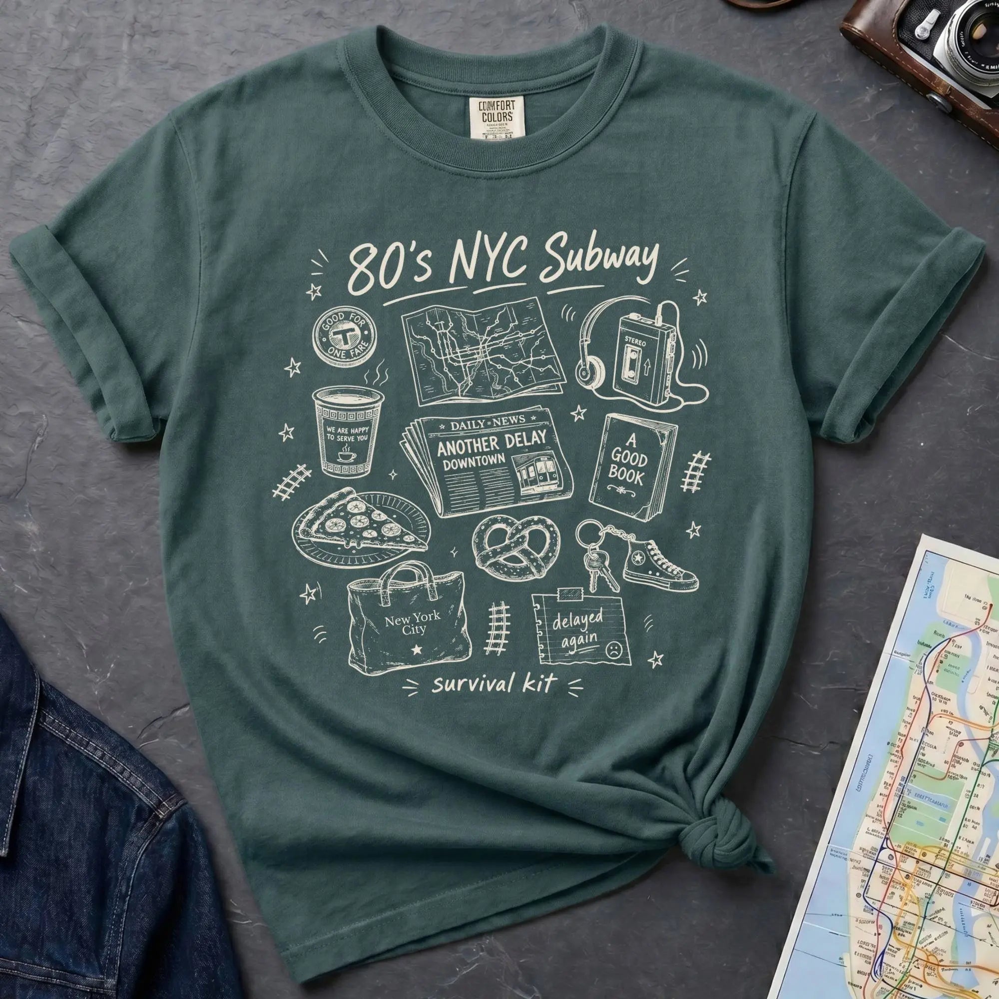 Blue spruce t-shirt with '80s NYC Subway Survival Kit' graphic design on a dark surface with a map and denim jacket.