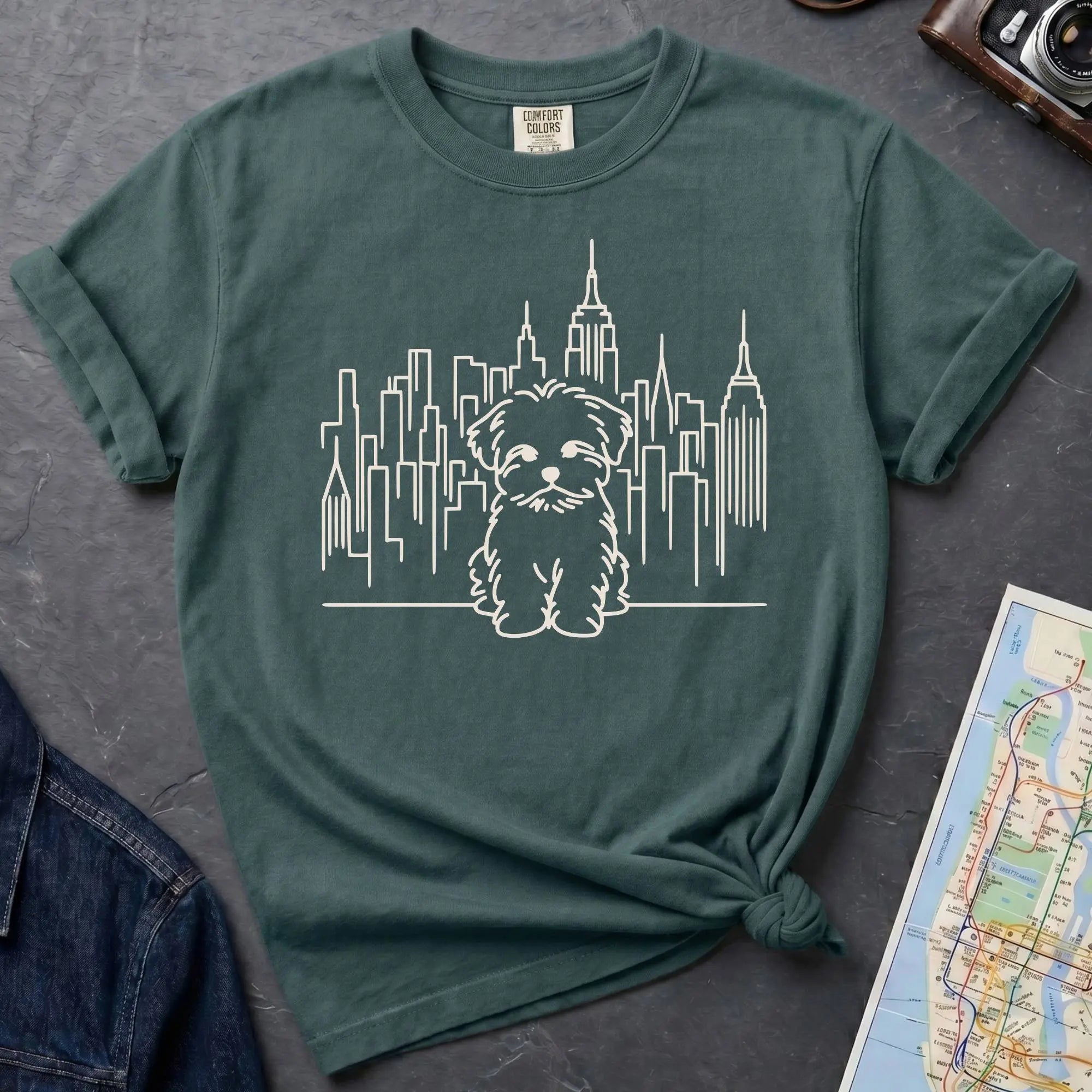 Blue Spruce t-shirt with a Maltese dog and cityscape design on a gray surface with a map and camera.