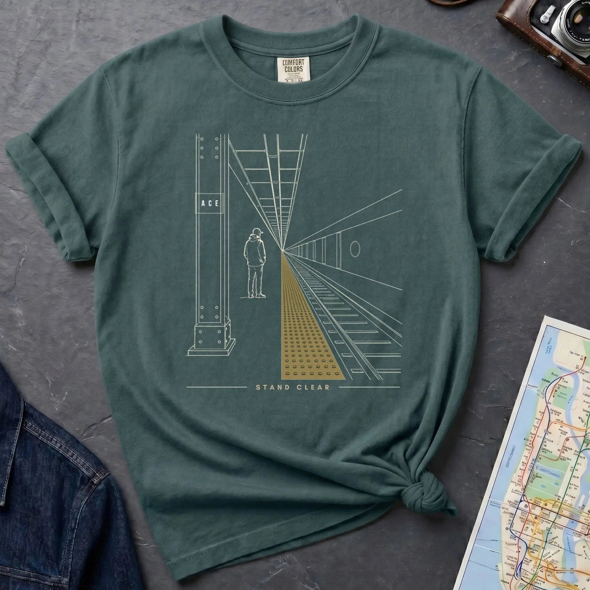 Blue Spruce Comfort Colors t-shirt featuring a minimalist NYC subway platform graphic with a lone figure, train tracks, and “Stand Clear” text.