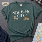 Blue spruce t-shirt with 'New York City' text and a roller skate and boom box on a dark surface with a map and camera.