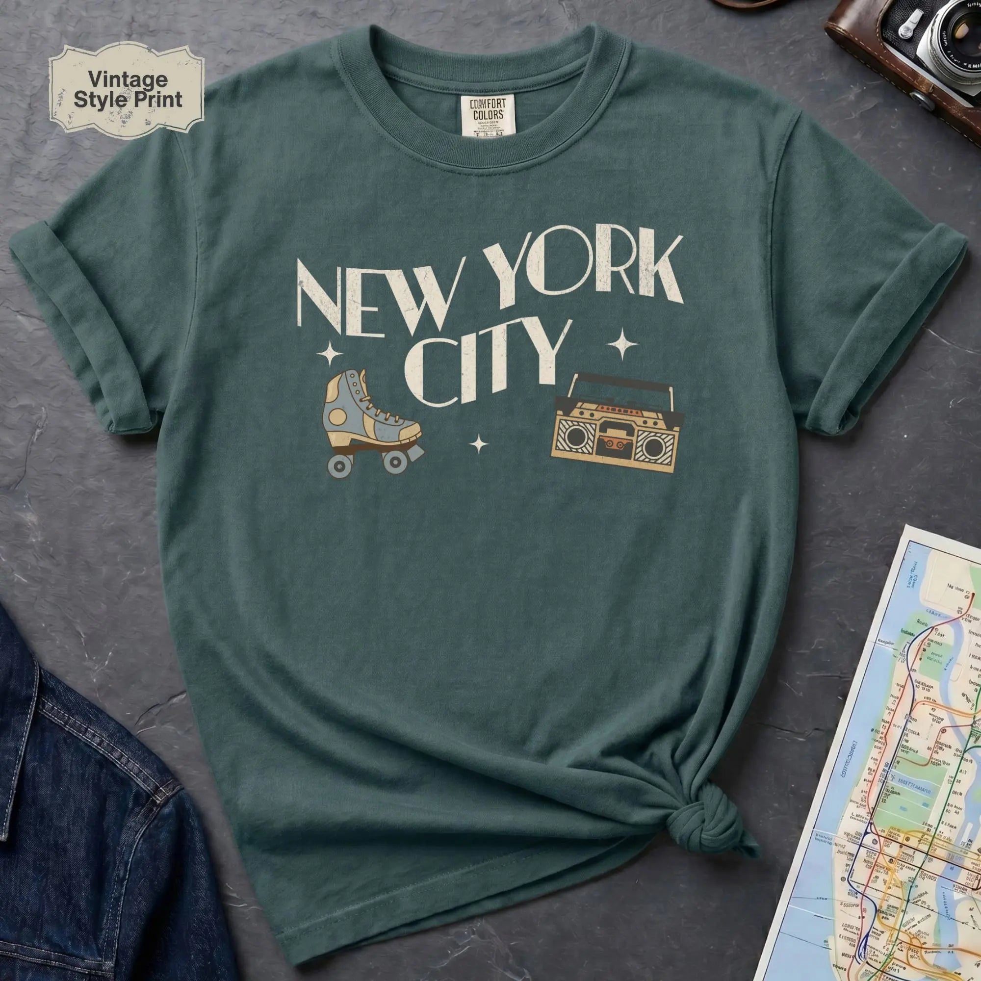 Blue spruce t-shirt with 'New York City' text and a roller skate and boom box on a dark surface with a map and camera.