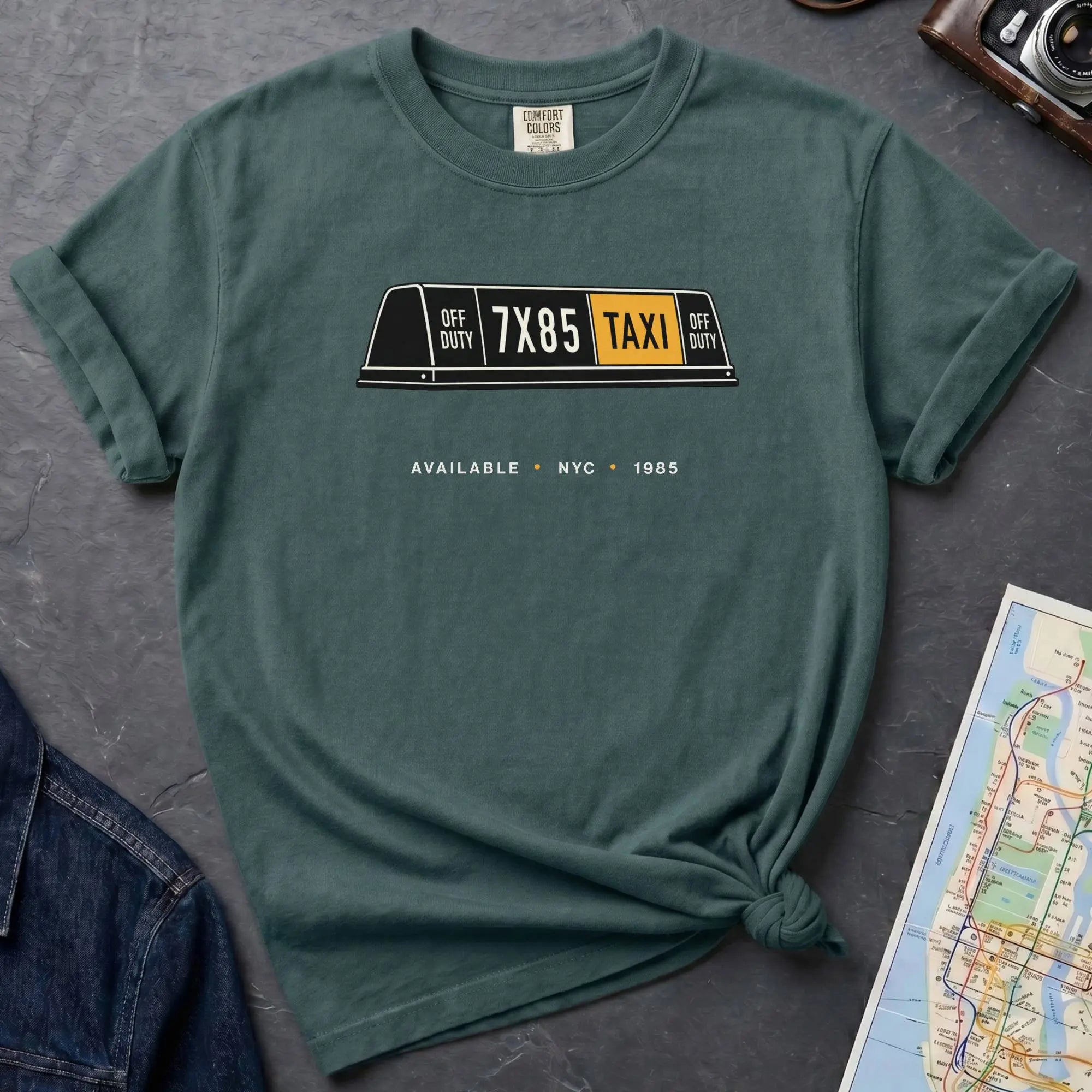 Blue Spruce Comfort Colors t-shirt featuring a vintage New York City taxi roof sign graphic with “Available • NYC • 1985” text.