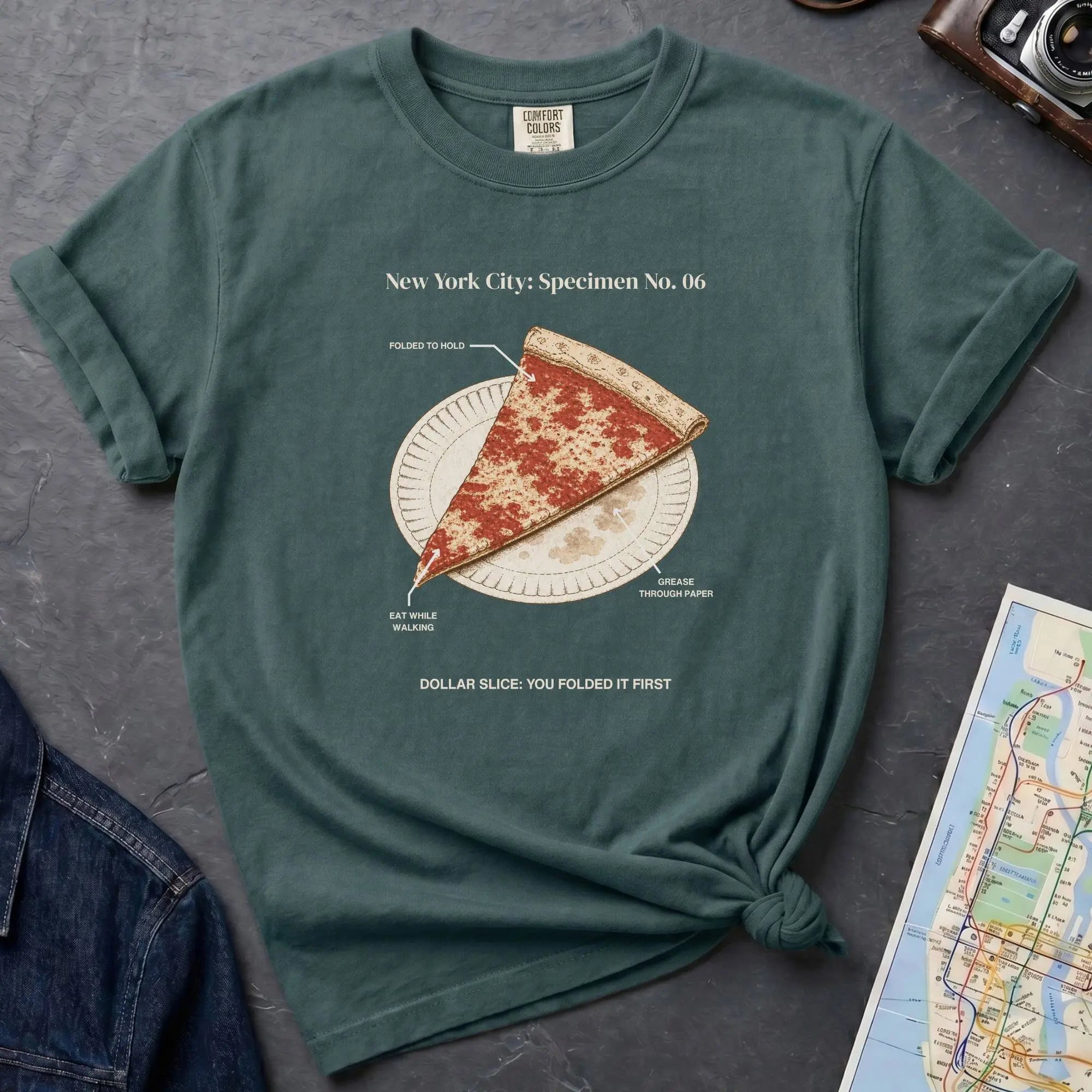 Blue spruce Comfort Colors t-shirt featuring a vintage New York City dollar slice pizza graphic with specimen-style callouts and a folded slice on a paper plate.