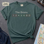 Blue Spruce t-shirt with 'The Bronx' and subway line numbers on a dark surface with a map and camera.