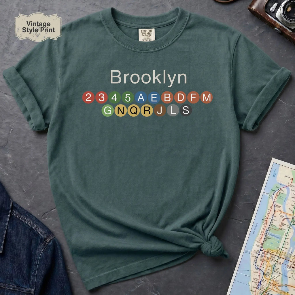 Blue Spruce t-shirt with 'Brooklyn' and subway line numbers on a dark surface with a map and camera.