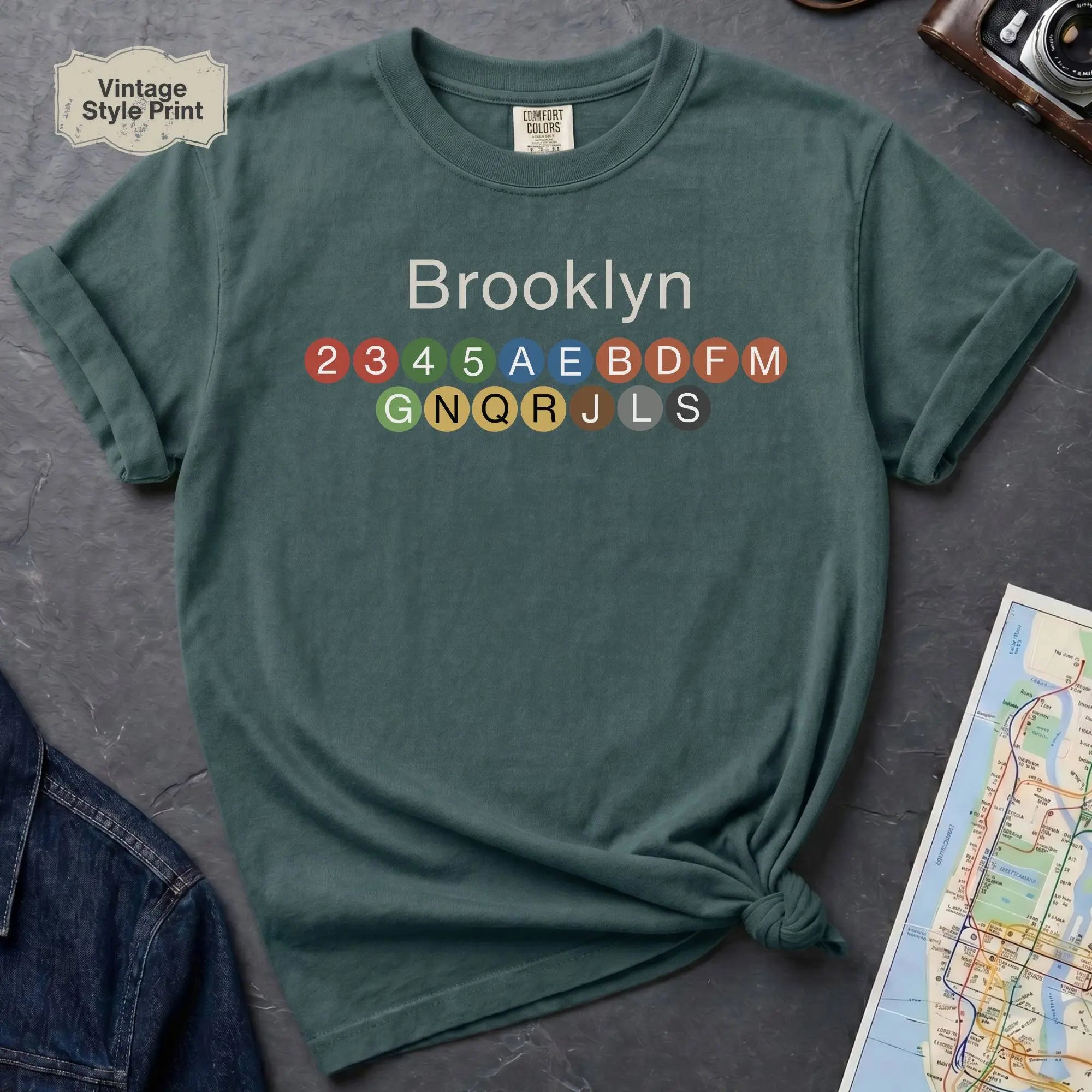 Blue Spruce t-shirt with 'Brooklyn' and subway line numbers on a dark surface with a map and camera.