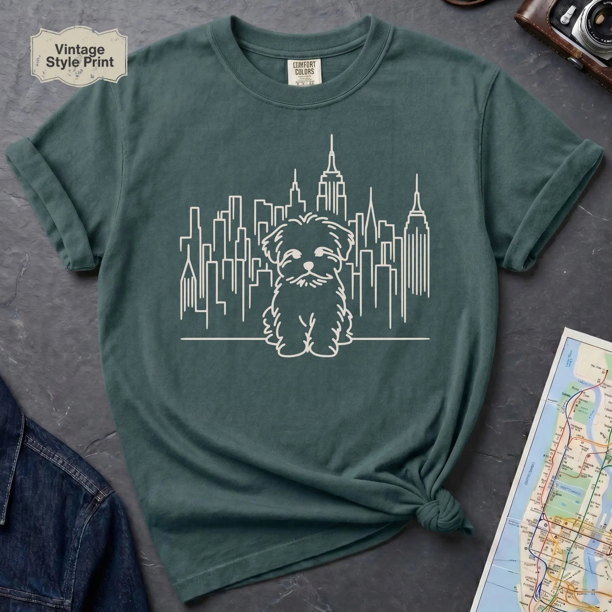 Blue Spruce t-shirt with a Maltese dog and cityscape design on a gray surface with a map and camera.