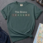 Blue spruce t-shirt with 'The Bronx' and subway line numbers on a dark surface with a map and camera.
