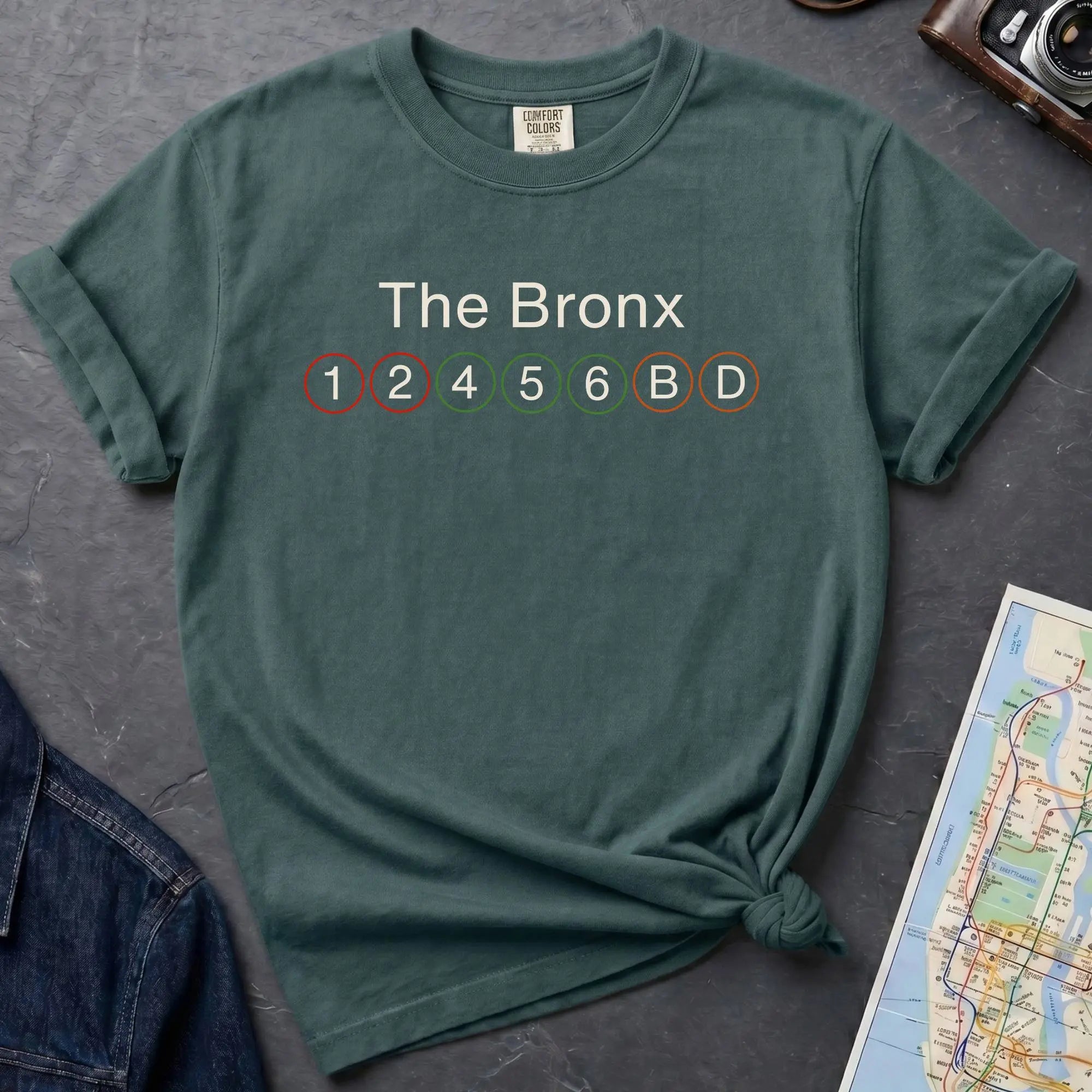 Blue spruce t-shirt with 'The Bronx' and subway line numbers on a dark surface with a map and camera.