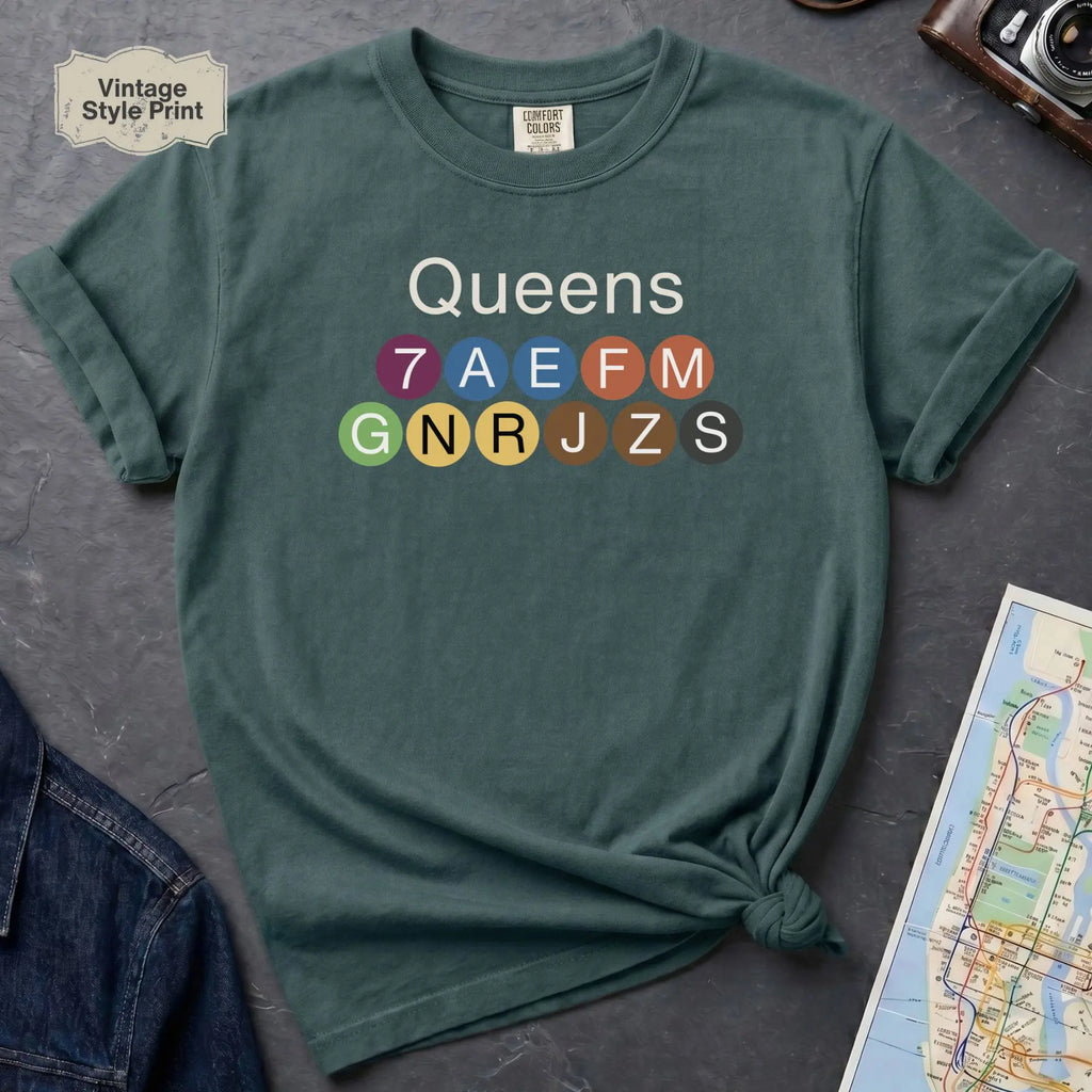 Blue spruce Black Comfort Colors t-shirt featuring a minimalist Queens subway line graphic with colorful NYC train icons.